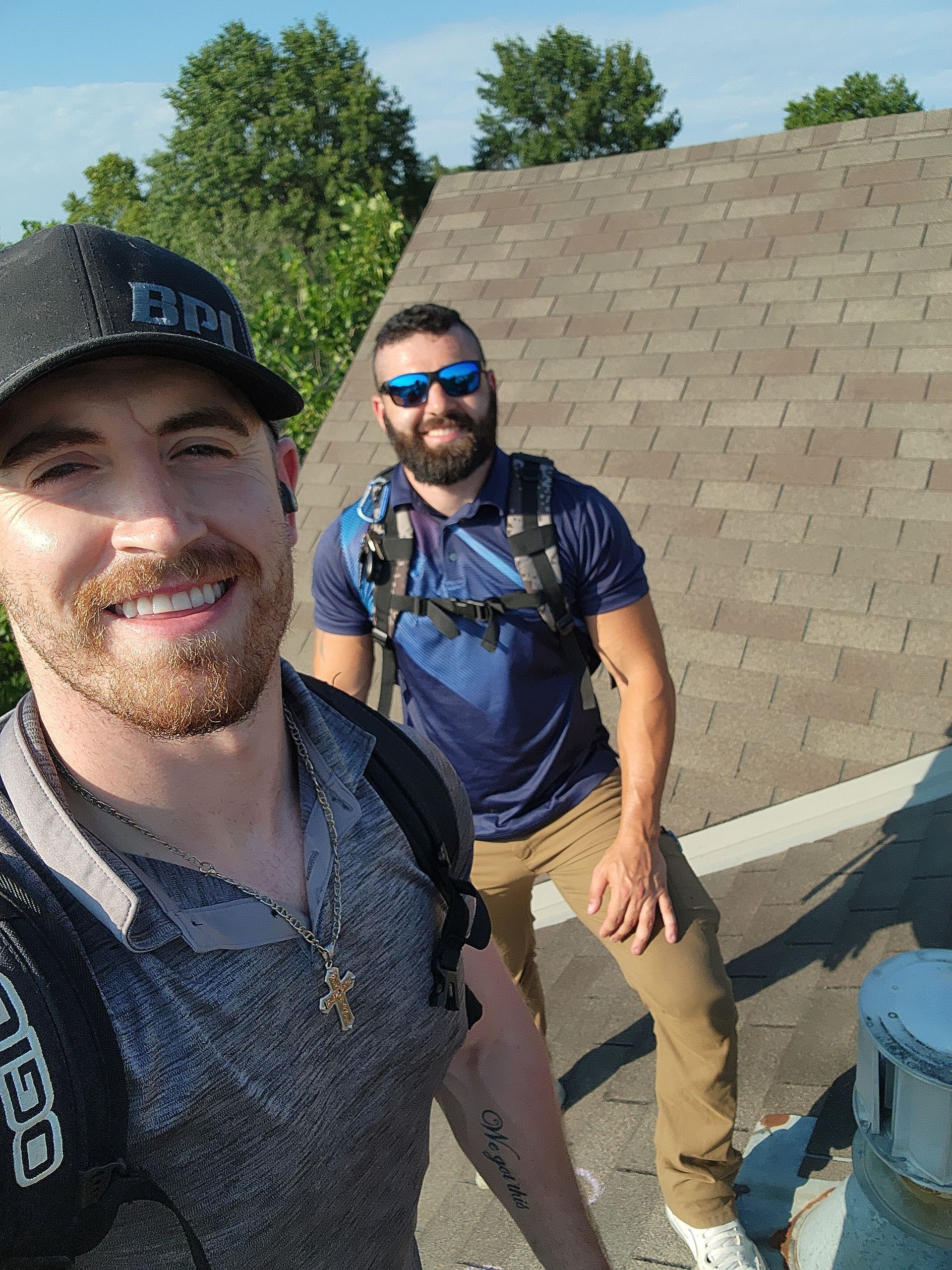 Two men on a rooftop, smiling. One takes a selfie; the other wears a backpack. Sunny day with trees in background.