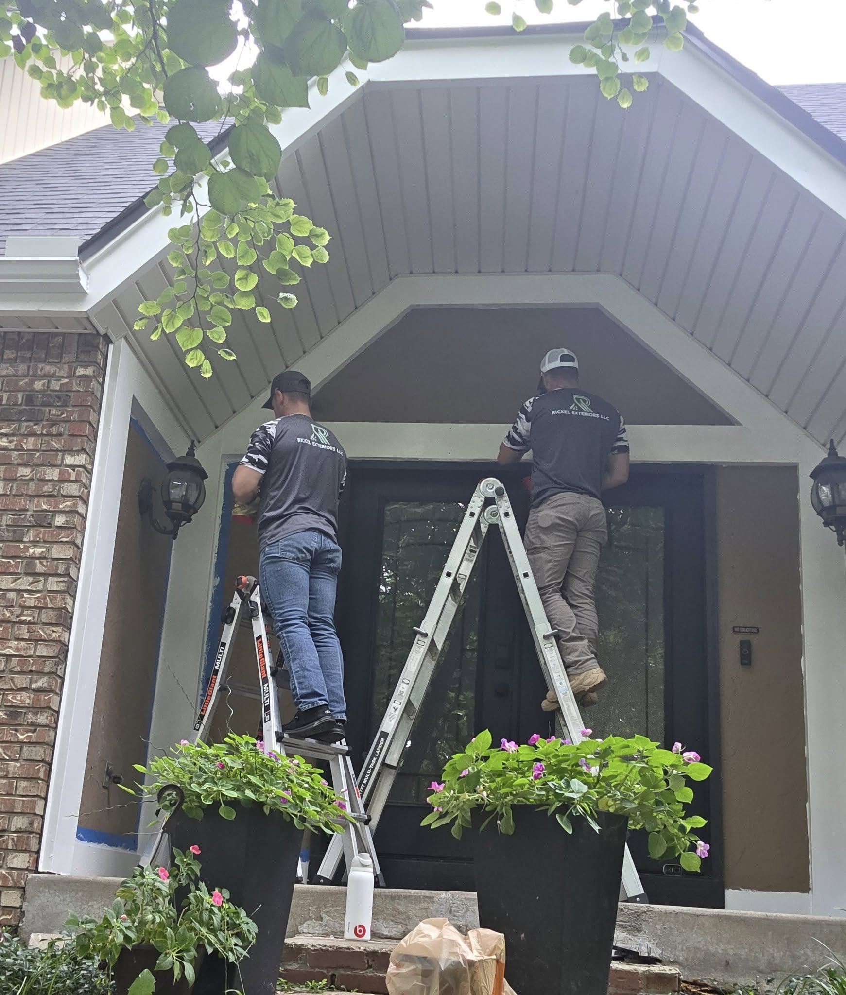 Two people on ladders painting the trim of a house's entrance. Black front door and potted plants are visible.