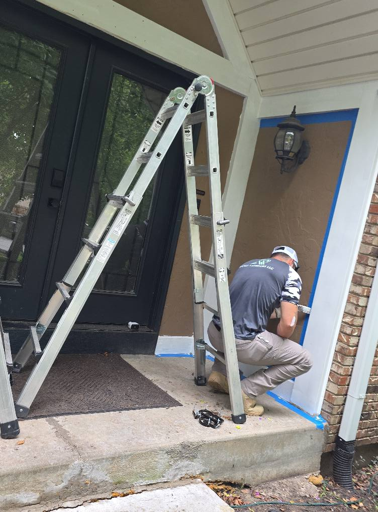 Person on a ladder painting exterior trim on a house. Blue tape protects adjacent surfaces; brown and white walls.