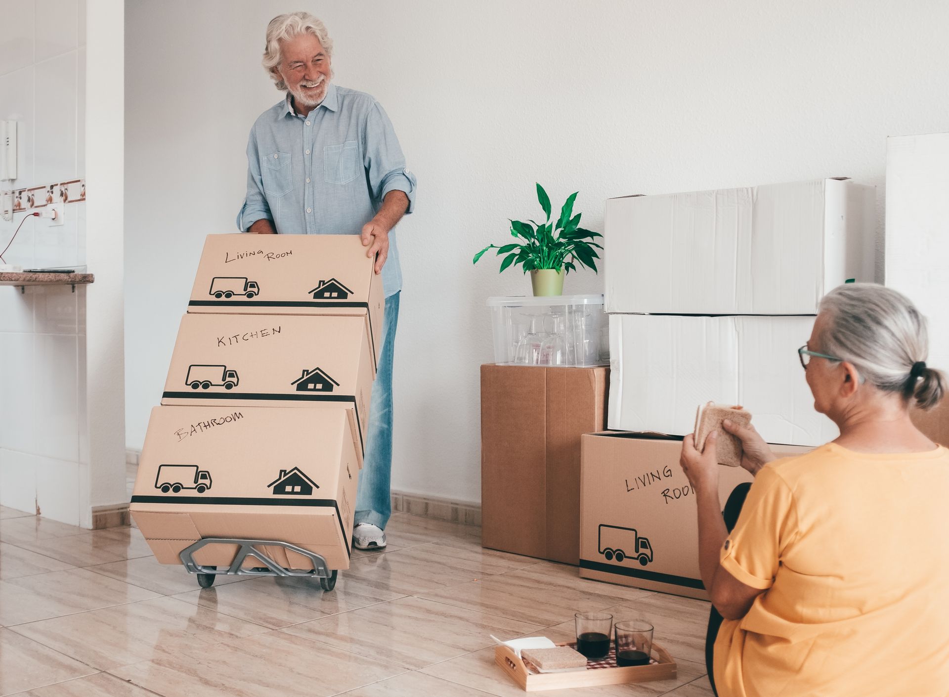Senior man pushing boxes on a cart, woman sitting and looking at a paper, inside a house.