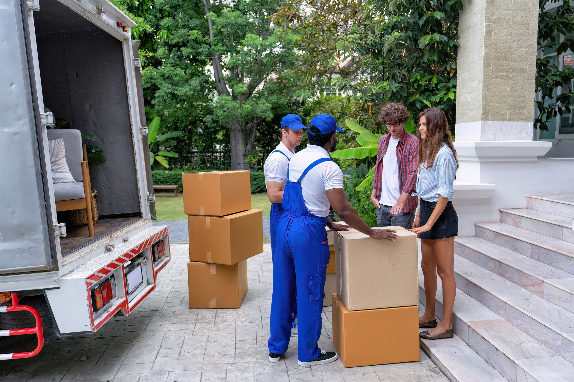 Movers unloading boxes from truck, talking to couple outside a house with steps.