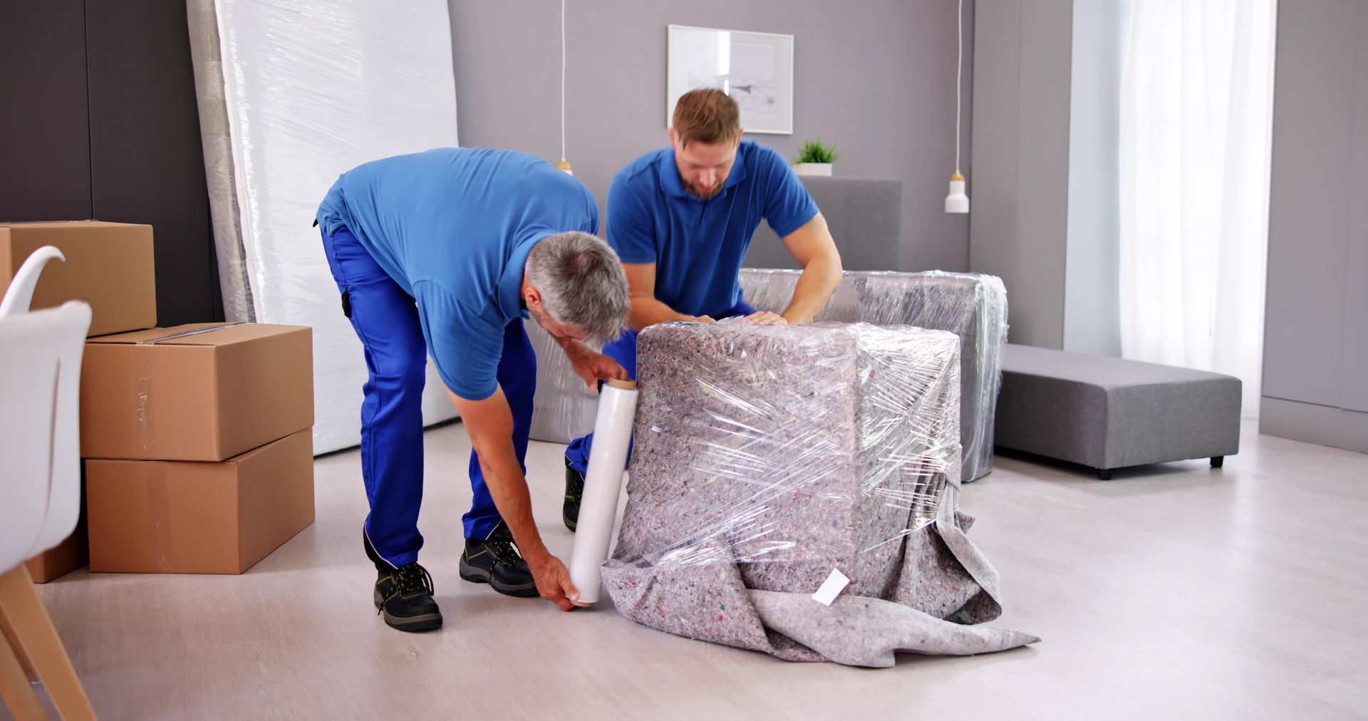 Two movers wrapping furniture in plastic wrap in a living room with boxes.