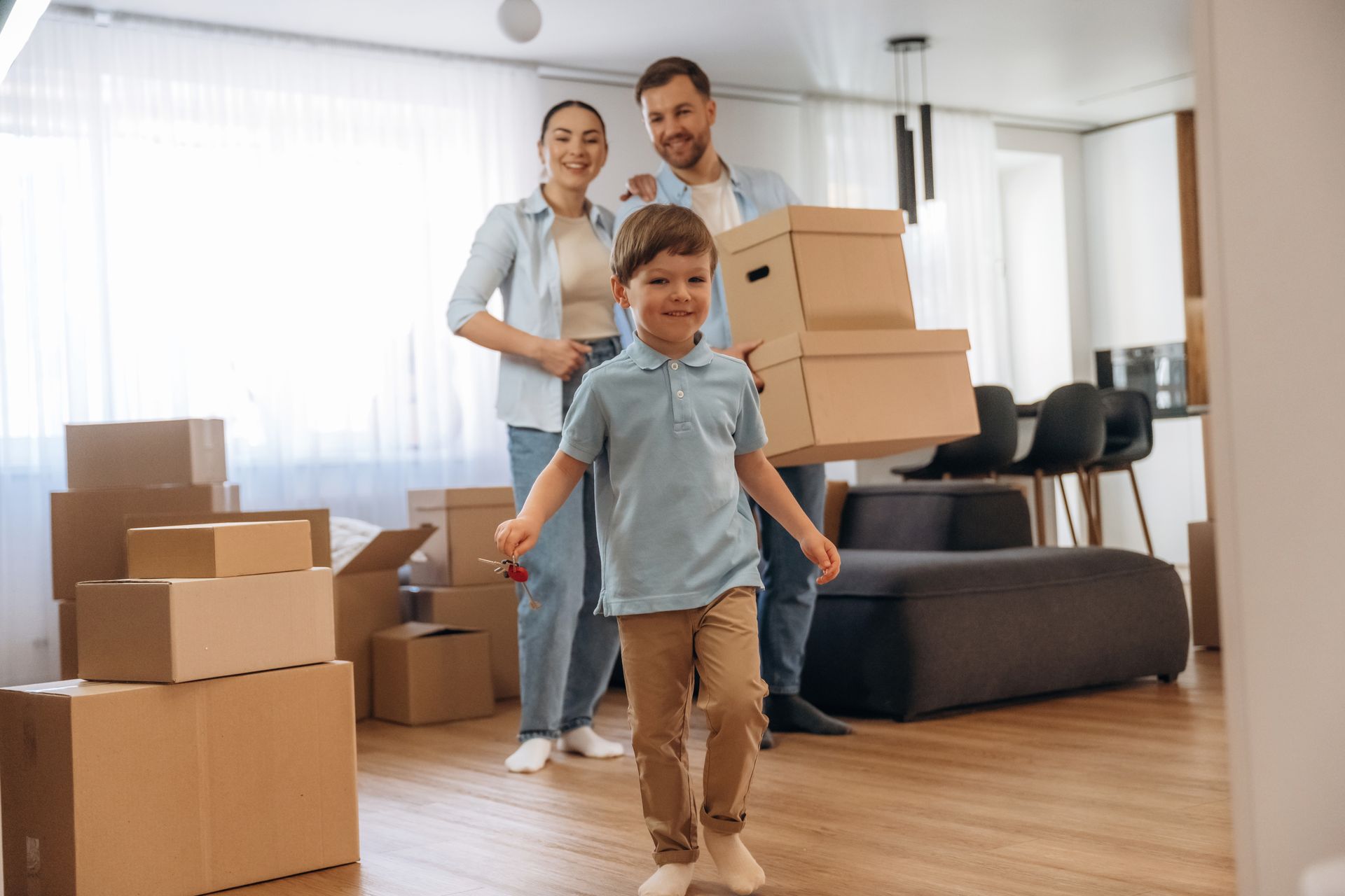 Family moving boxes into a new home. Boy runs, parents smile carrying boxes.