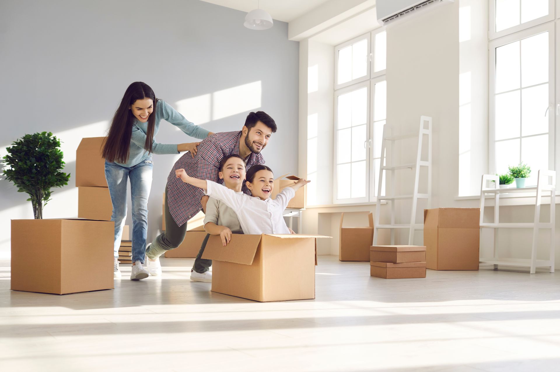 Family playing in a new home; children in a box being pushed by parents near moving boxes.