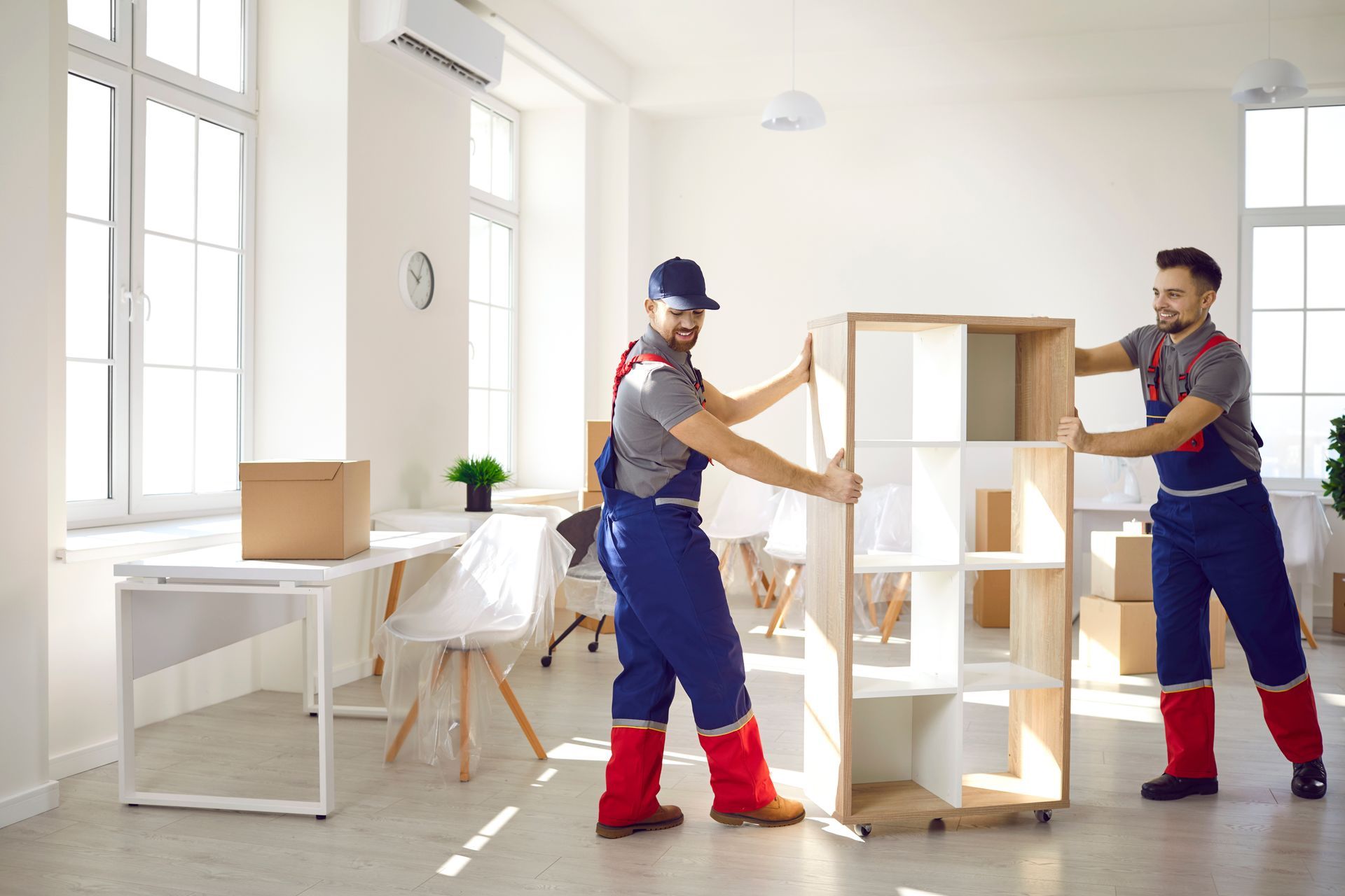 Two movers in blue coveralls and red boots carry a wooden shelf in a bright office.