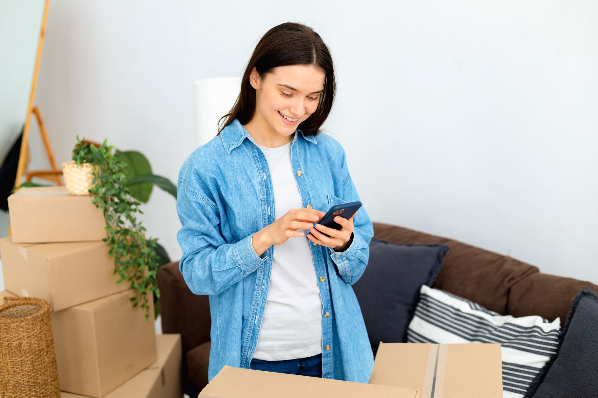 Woman in denim shirt smiles, looking at phone in front of cardboard boxes in room.