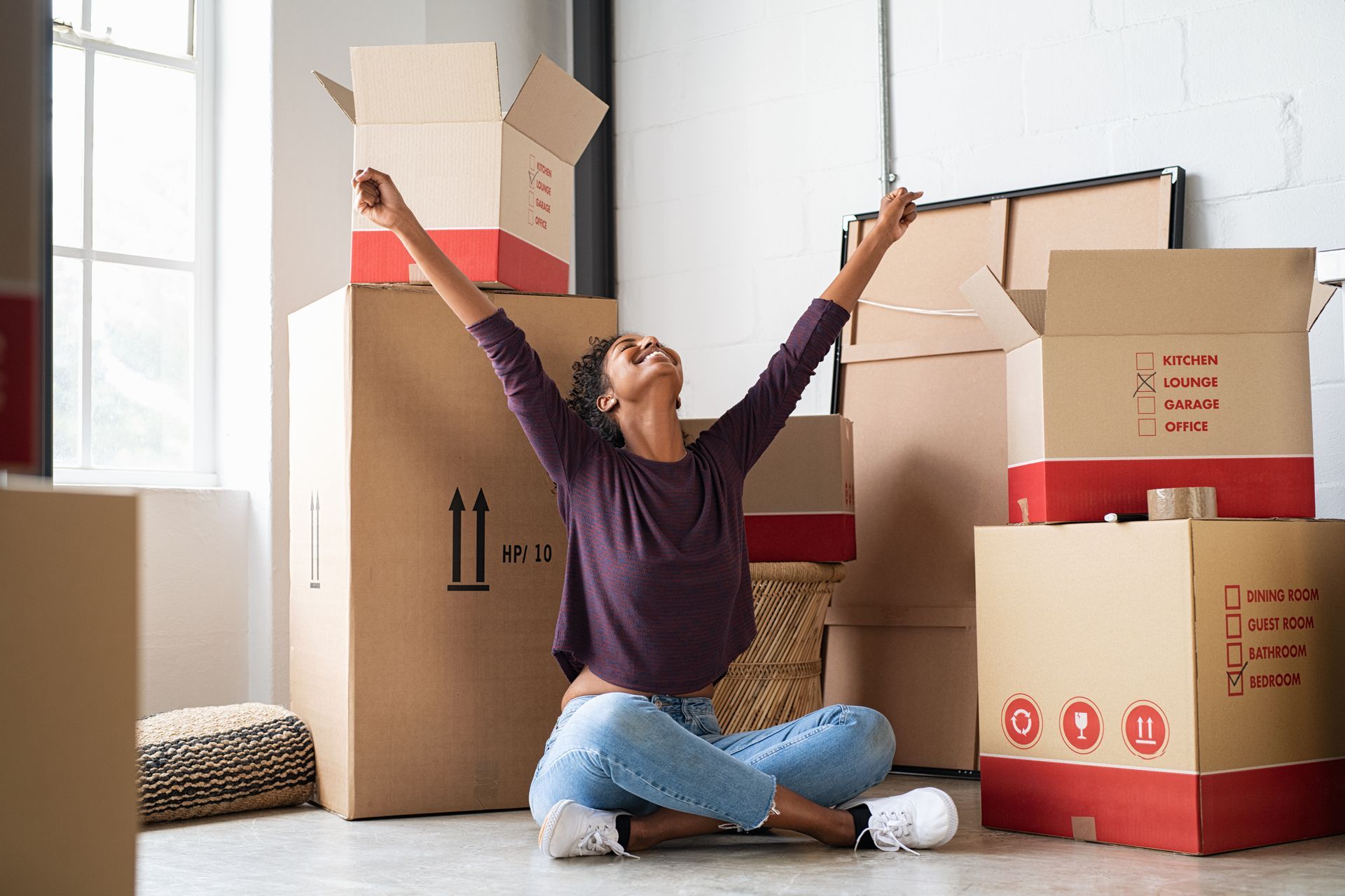 Woman in jeans sits amidst moving boxes, arms raised in triumph.