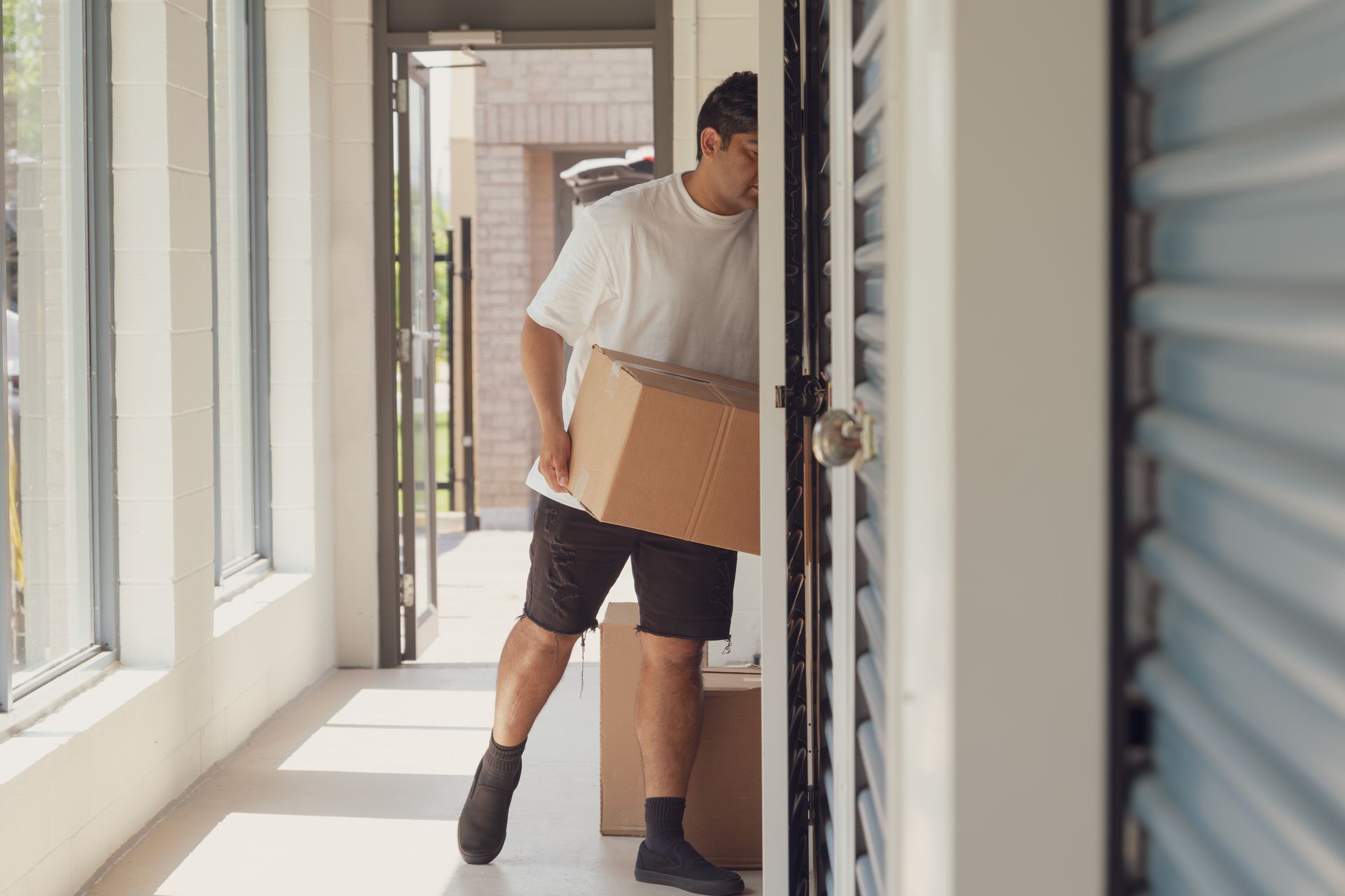 Man carrying a cardboard box through a doorway, likely moving. Bright hallway with sunlight.