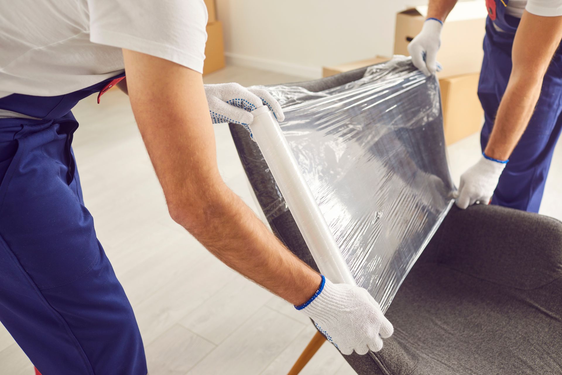 Two movers in blue coveralls wrap a gray chair with plastic.