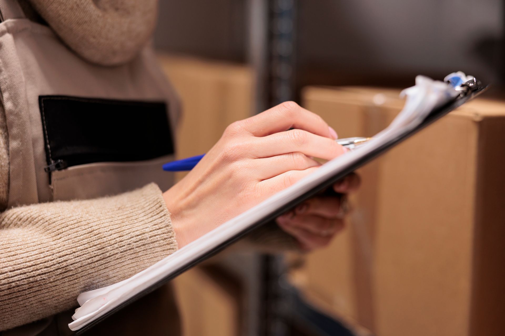 Person in a warehouse writing on a clipboard with a pen. Boxes are visible in the background.