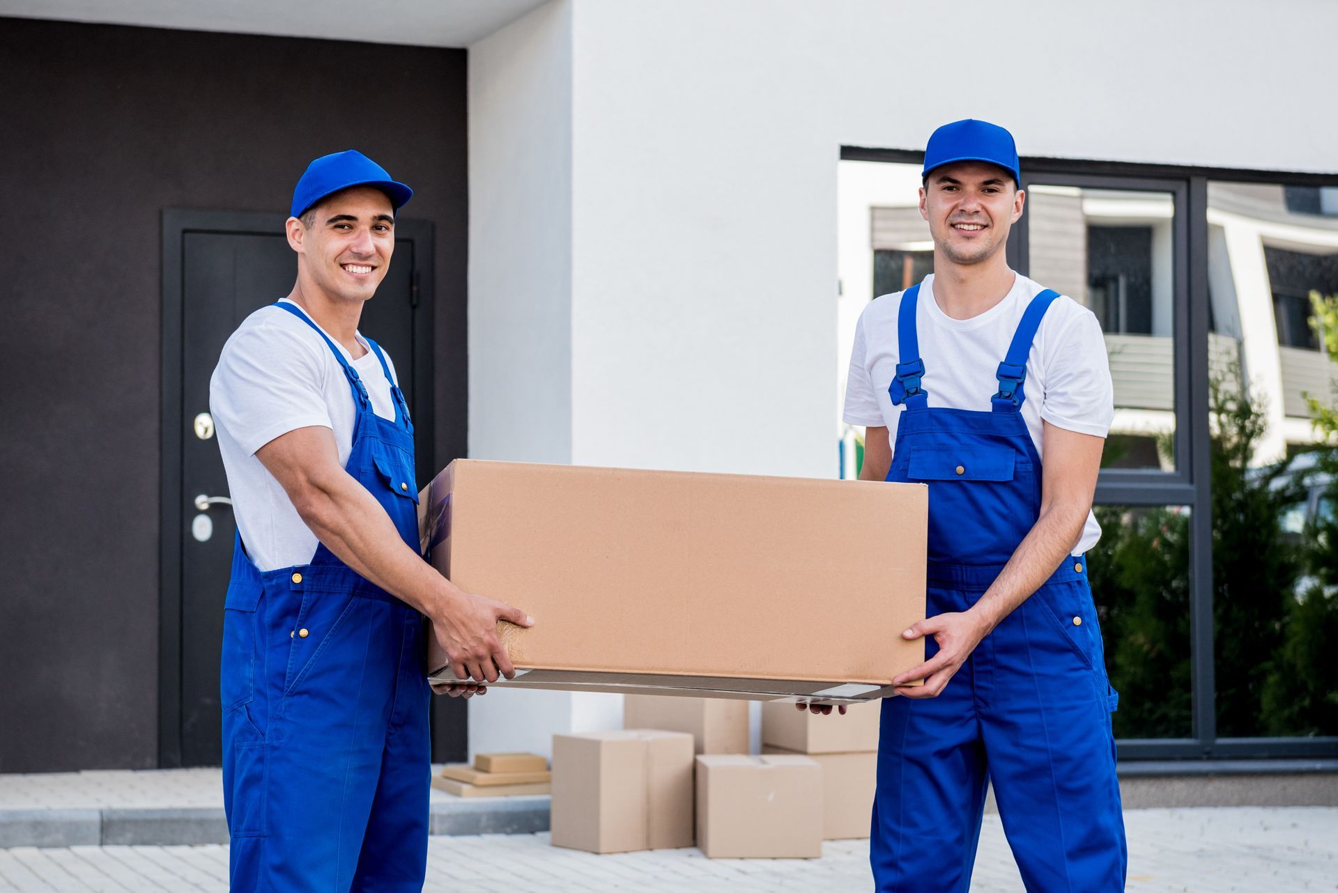 Two movers are carrying a cardboard box in front of a house.