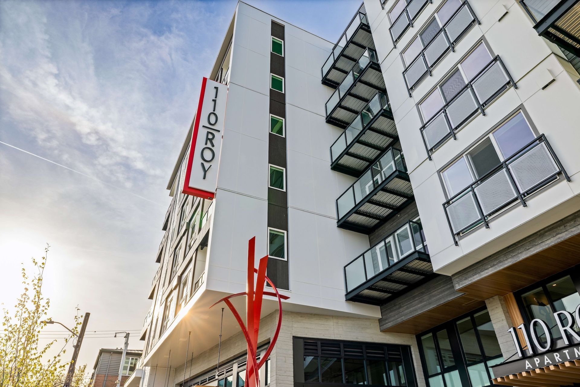 Modern apartment building, 110 Roy, with balconies and a red sign, sunny day.