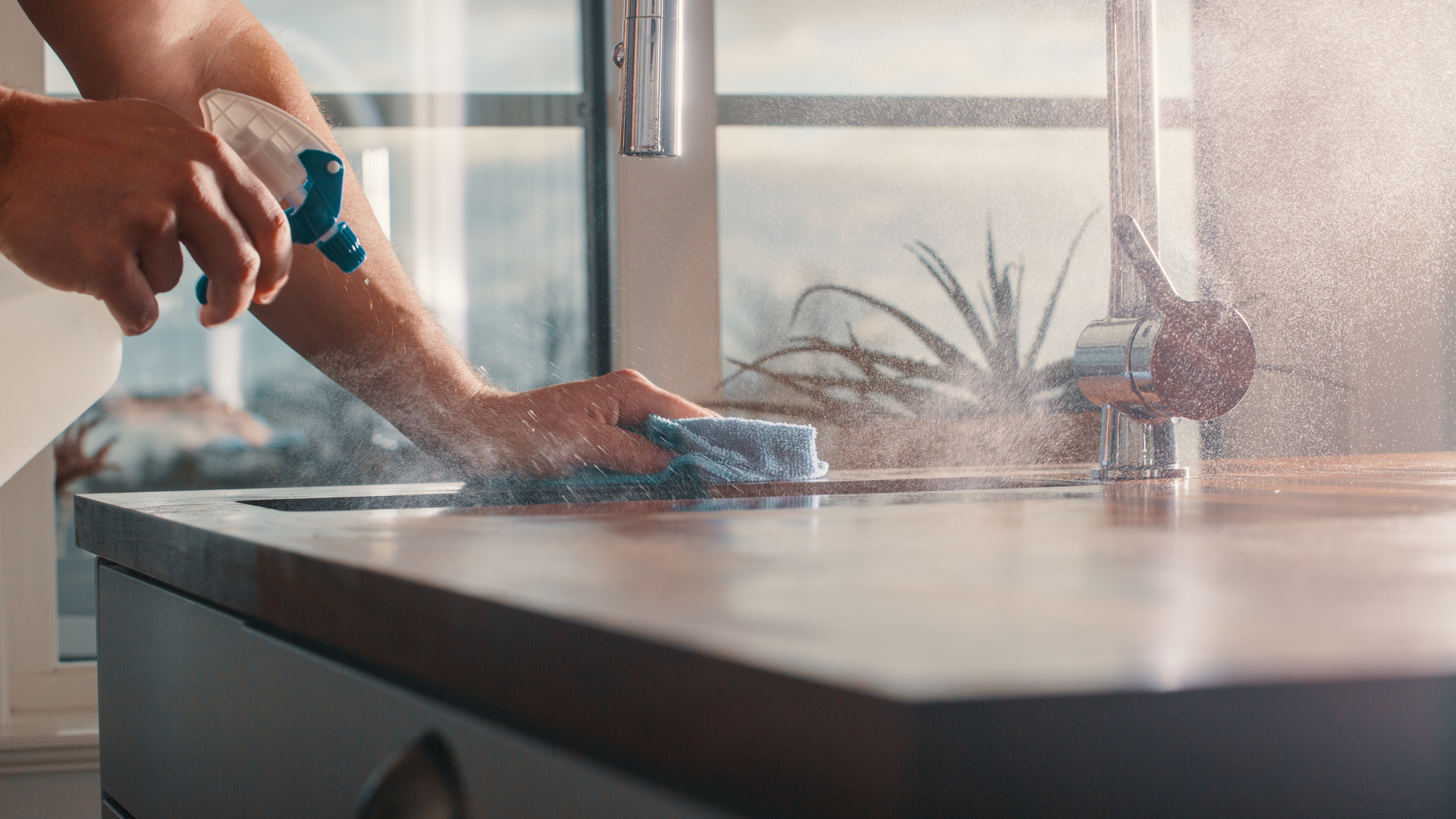 Person cleaning a countertop with spray bottle and cloth in a kitchen, near a window.