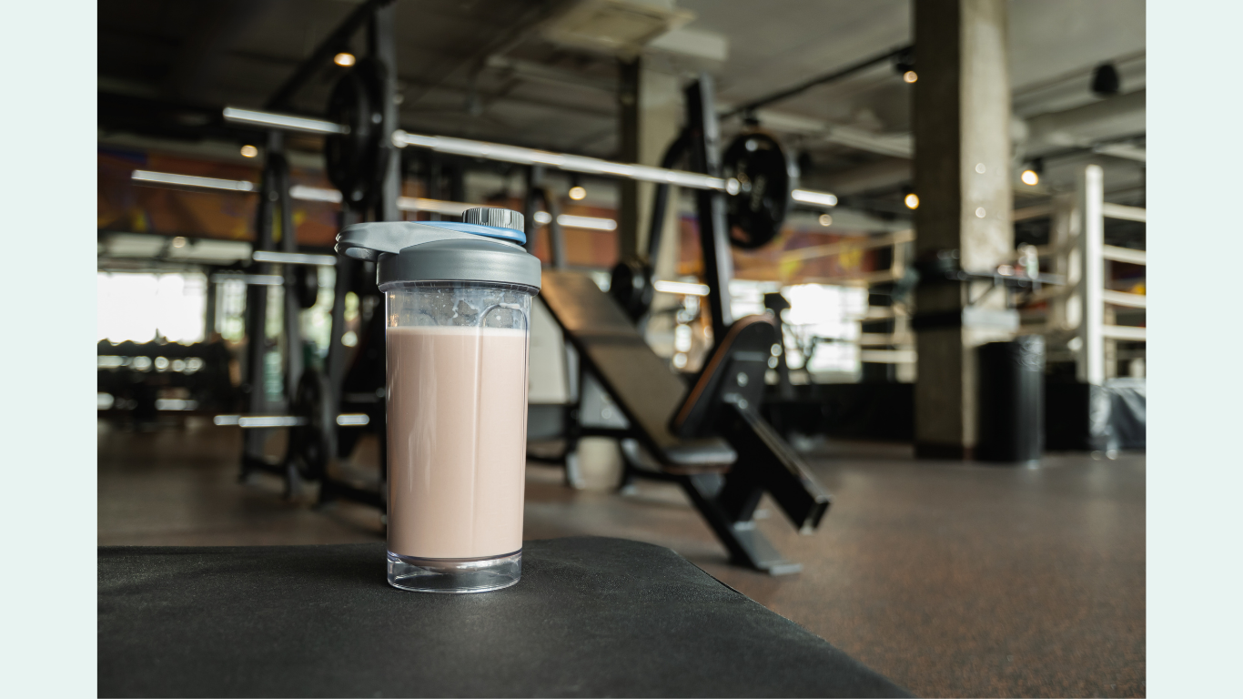 A protein shake in a clear tumbler sits on a mat in a gym