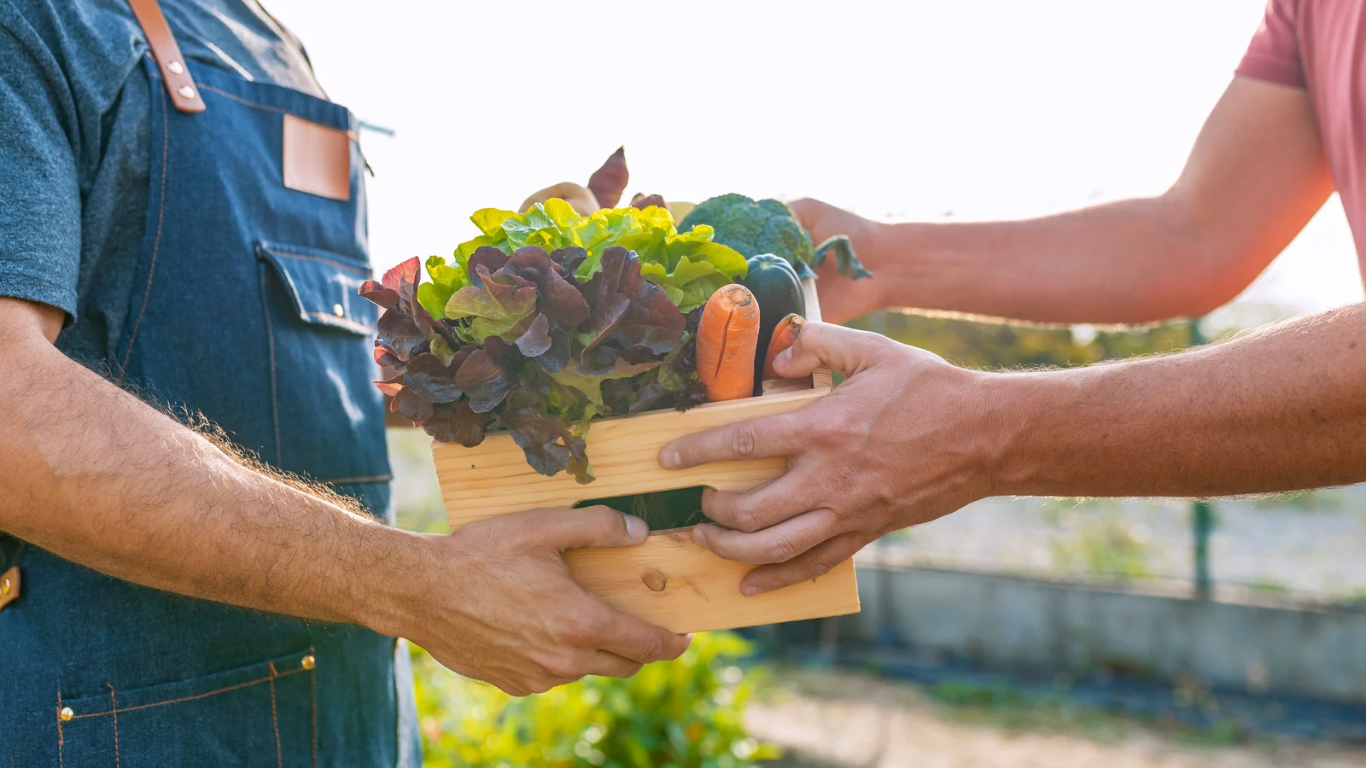 A person hands another person a wooden crate full of locally-sourced produce.