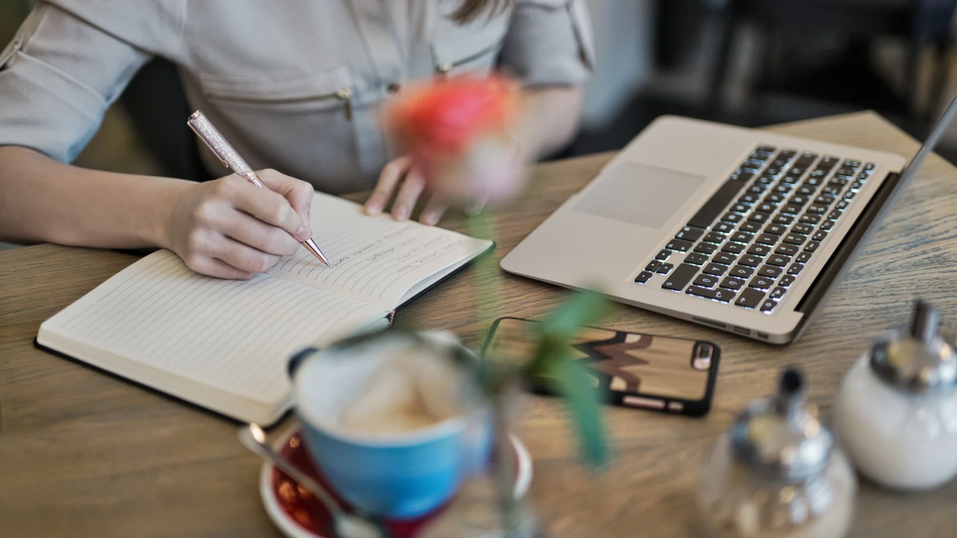 A woman from the chest down writes in a journal on a wooden desk with a labtop, coffee cup, & phone