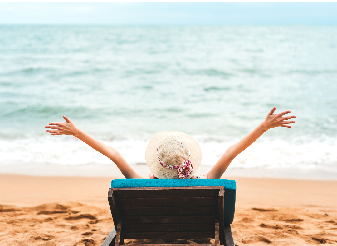 A woman in a straw hat sitting beachside in a reclining chair with rreaches her arms upwards