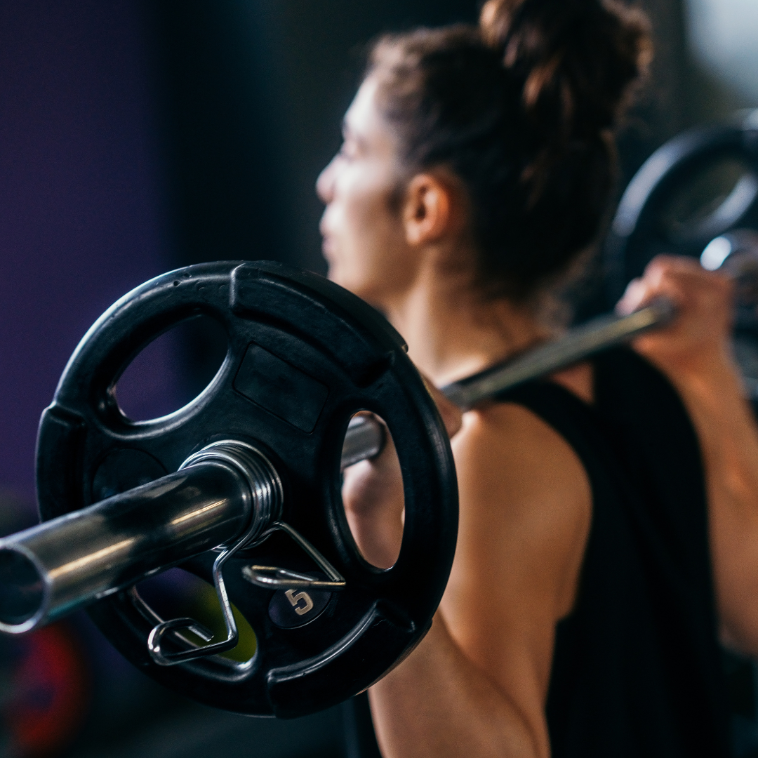 A woman is with her hair up in a bun with a black tank top is facing away, lifting a barbell