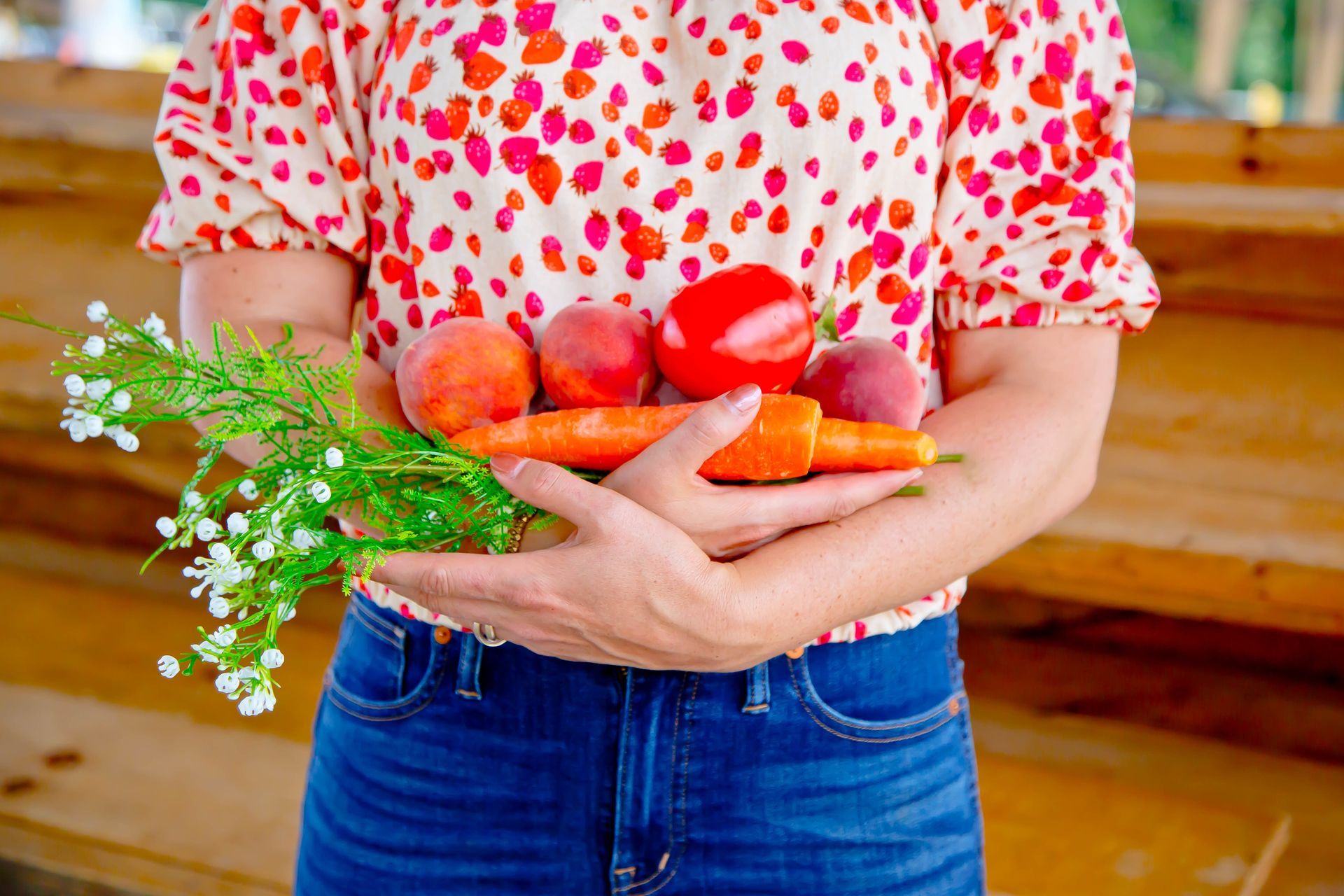 A woman's arms holding produce in front of her, including carrots, peppers and apples.