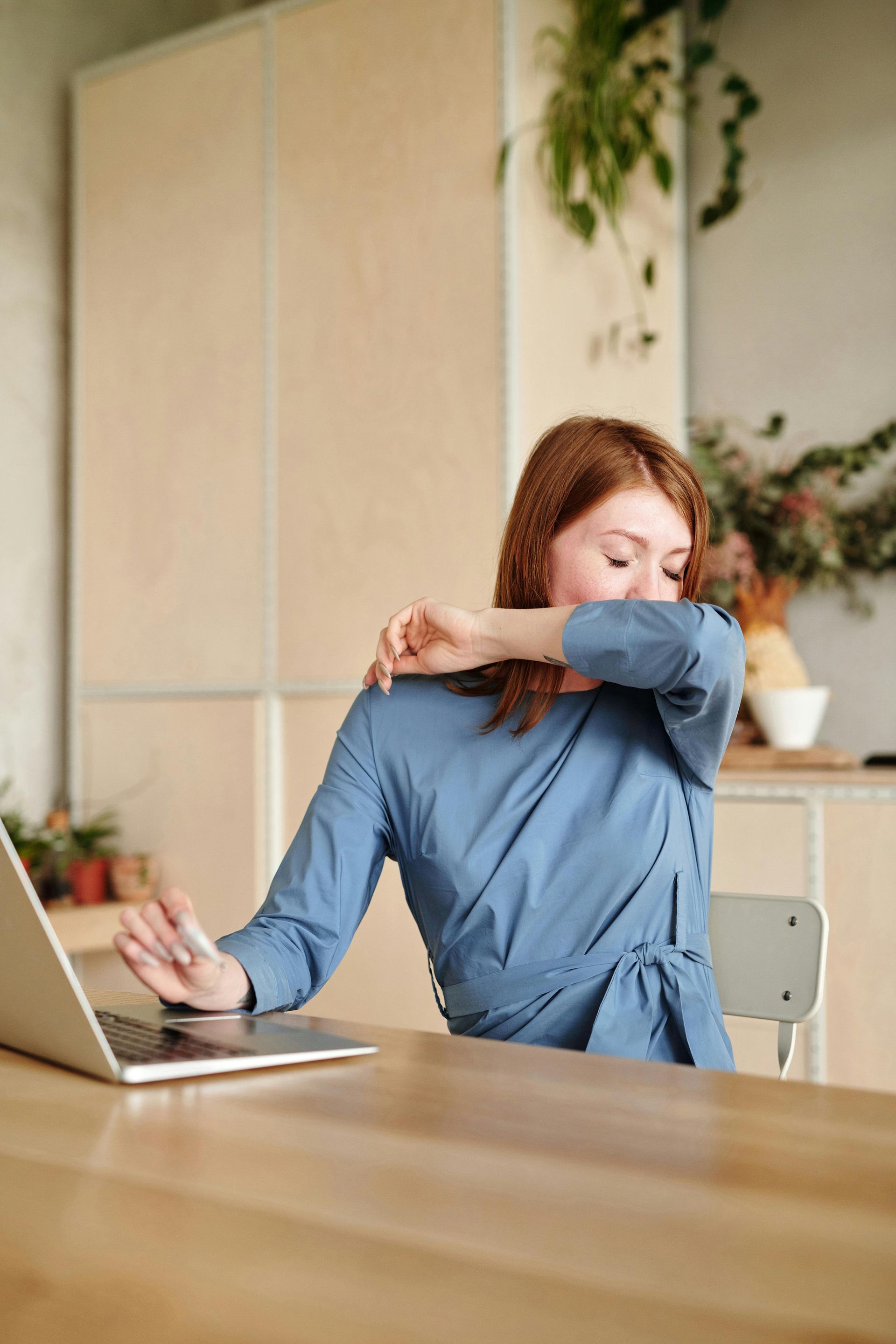 A woman is sitting at a table with a laptop and coughing into her hand.