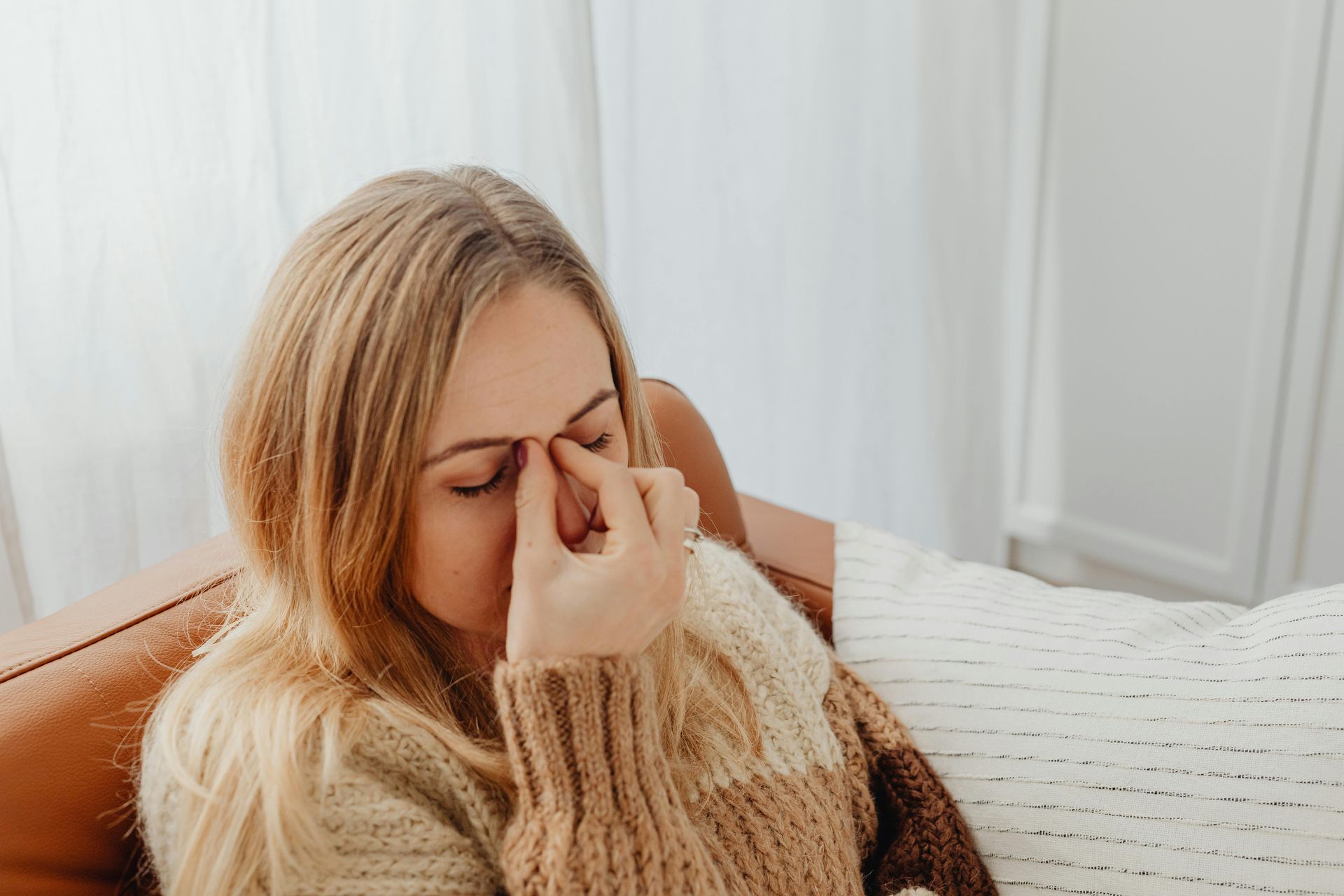 A woman is sitting on a couch holding her nose because she has a headache.