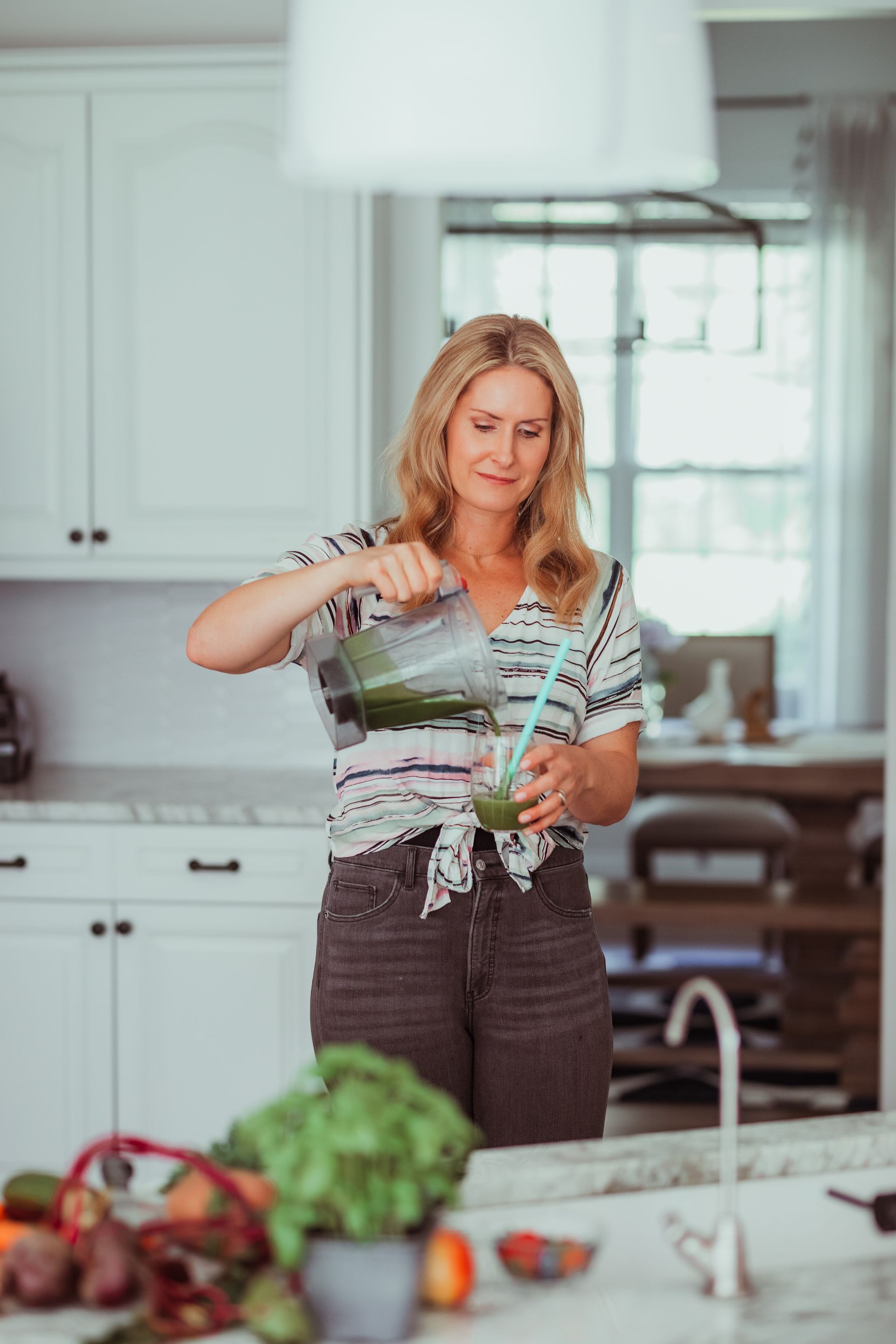 A woman is pouring a green smoothie into a glass in a kitchen.