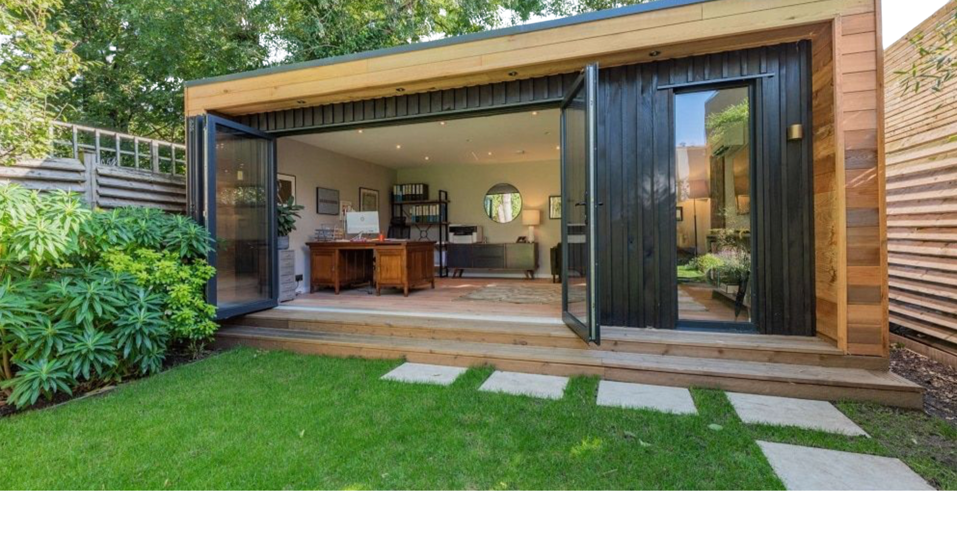 Backyard office with open glass doors, wooden deck, green grass, and garden.