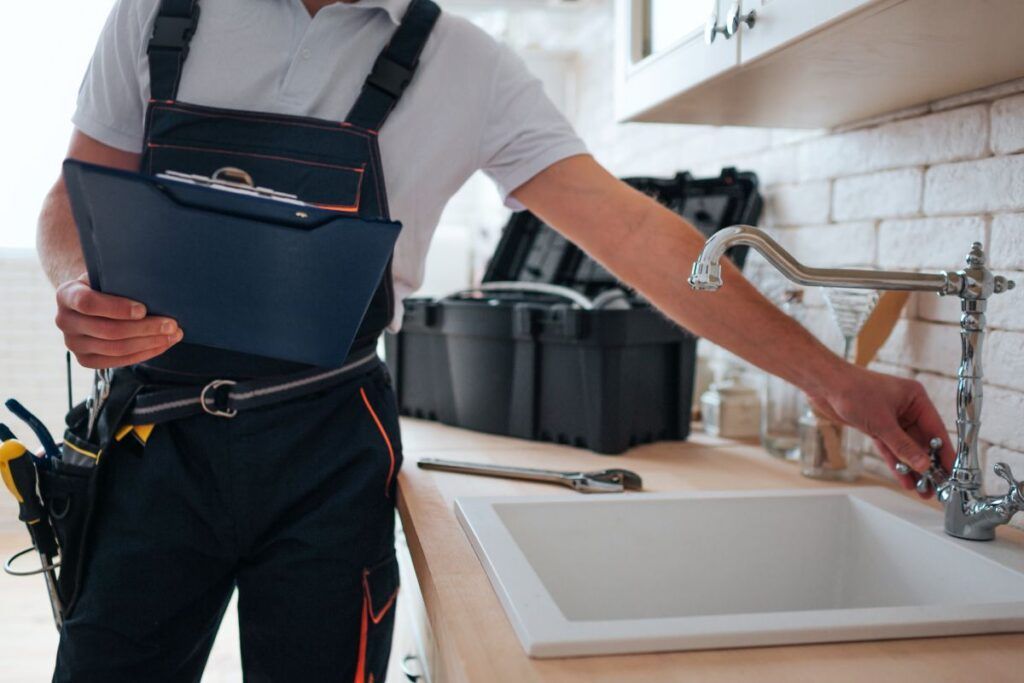 A plumber is fixing a faucet in a kitchen while holding a clipboard.