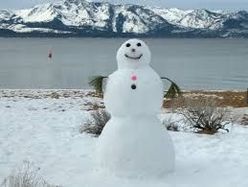 A snowman is standing in the snow near a lake with mountains in the background.