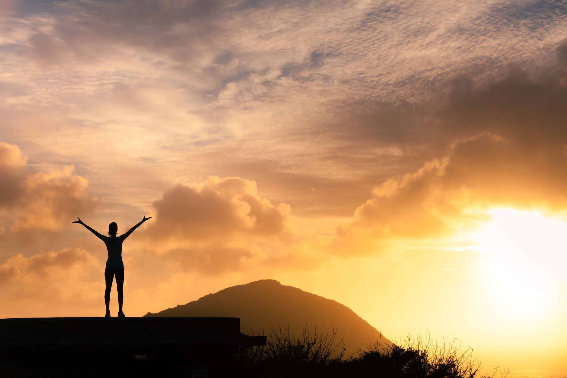Person with arms raised stands on a platform, silhouetted against a sunrise over a mountain.