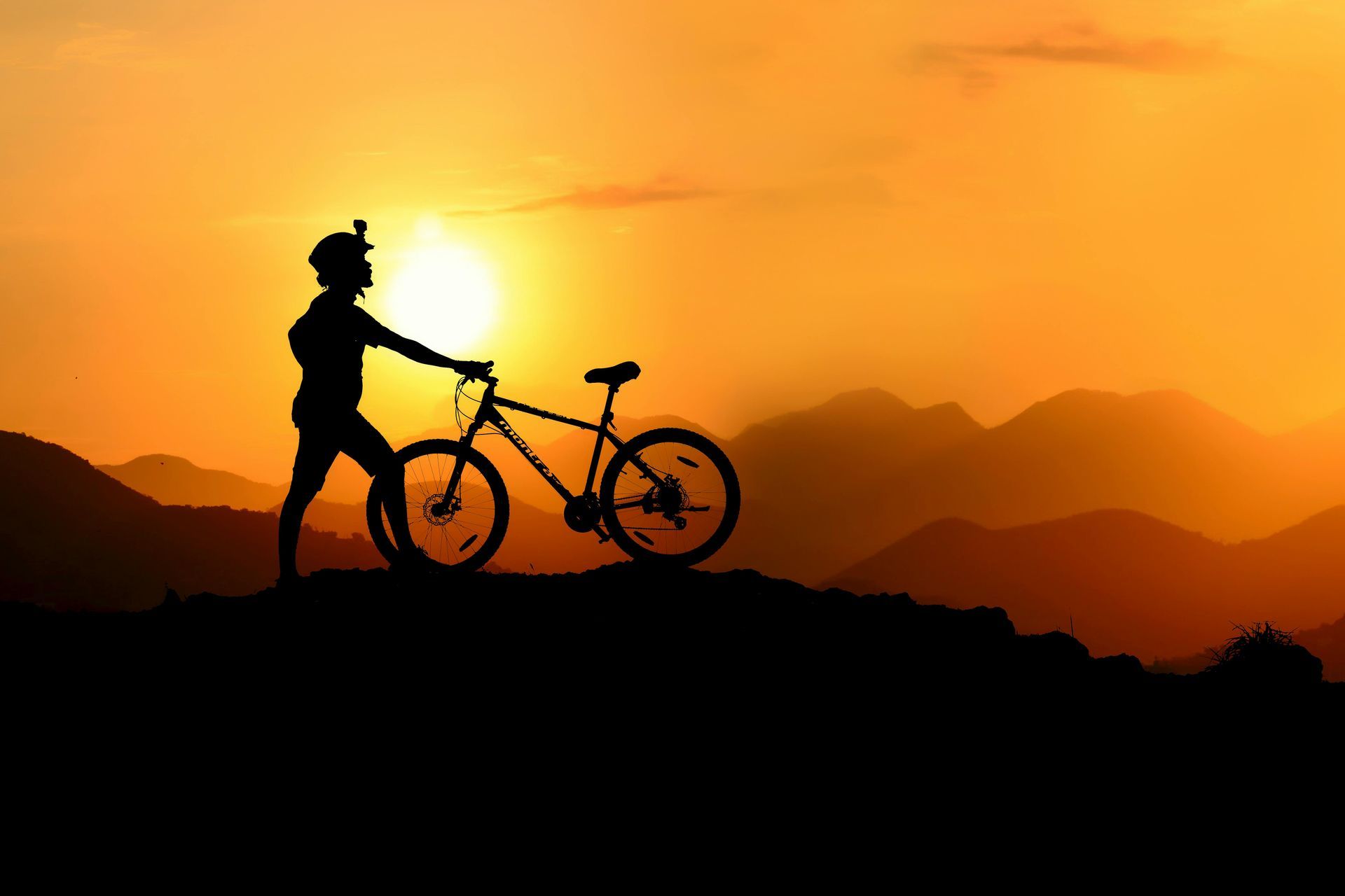 Silhouette of person with bike on mountaintop at sunset.