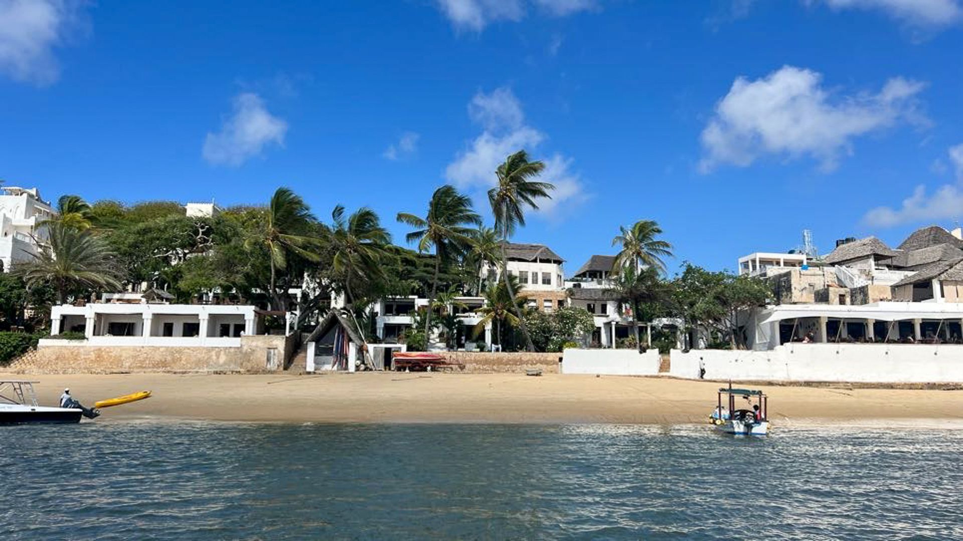 A boat is floating on the water near a beach with buildings in the background.