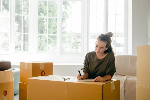 Une femme est assise sur une boîte en carton dans un salon et écrit sur un morceau de papier.