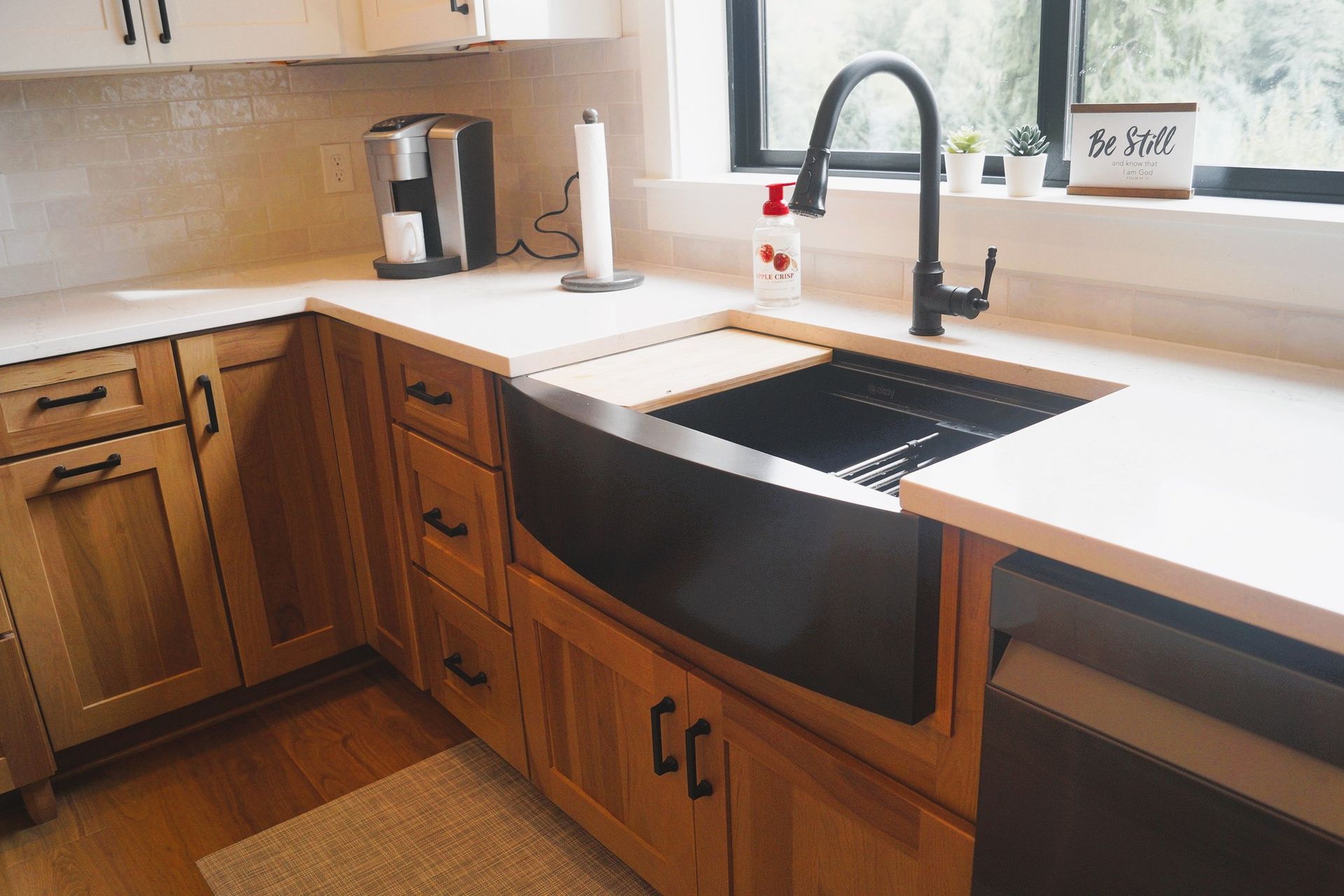 Sunlit kitchen with wooden cabinets, black sink, white countertops, and a window over the sink