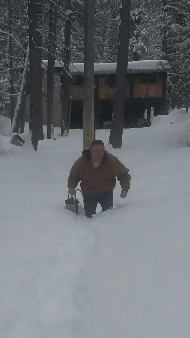 A person in a brown coat walks through deep snow in a wooded area toward a house in the background.