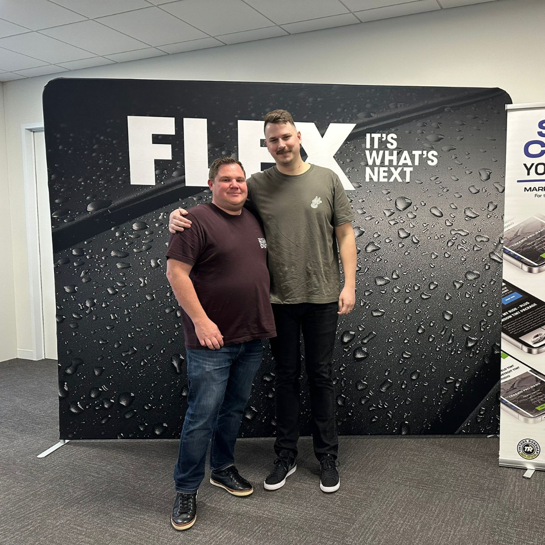 Two men are posing for a picture in front of a sign that says flex it 's what 's next.