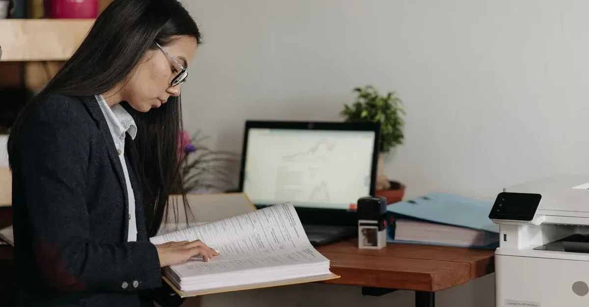 Woman in glasses reviewing documents at desk with laptop, printer, and office supplies for car detailing software guide.