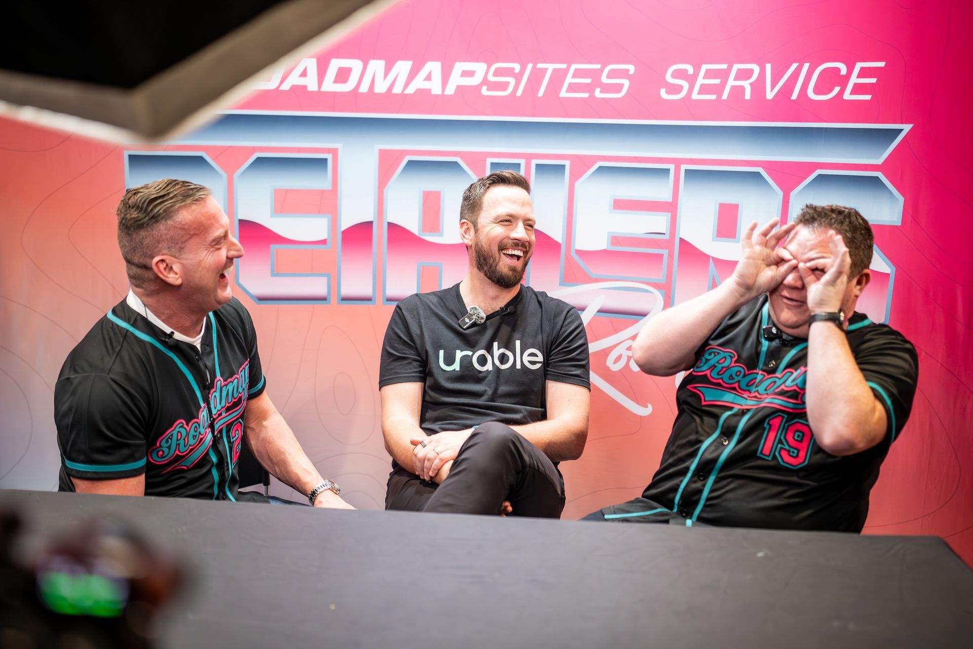 Three men laughing, one making glasses with fingers, in front of a pink backdrop with text.