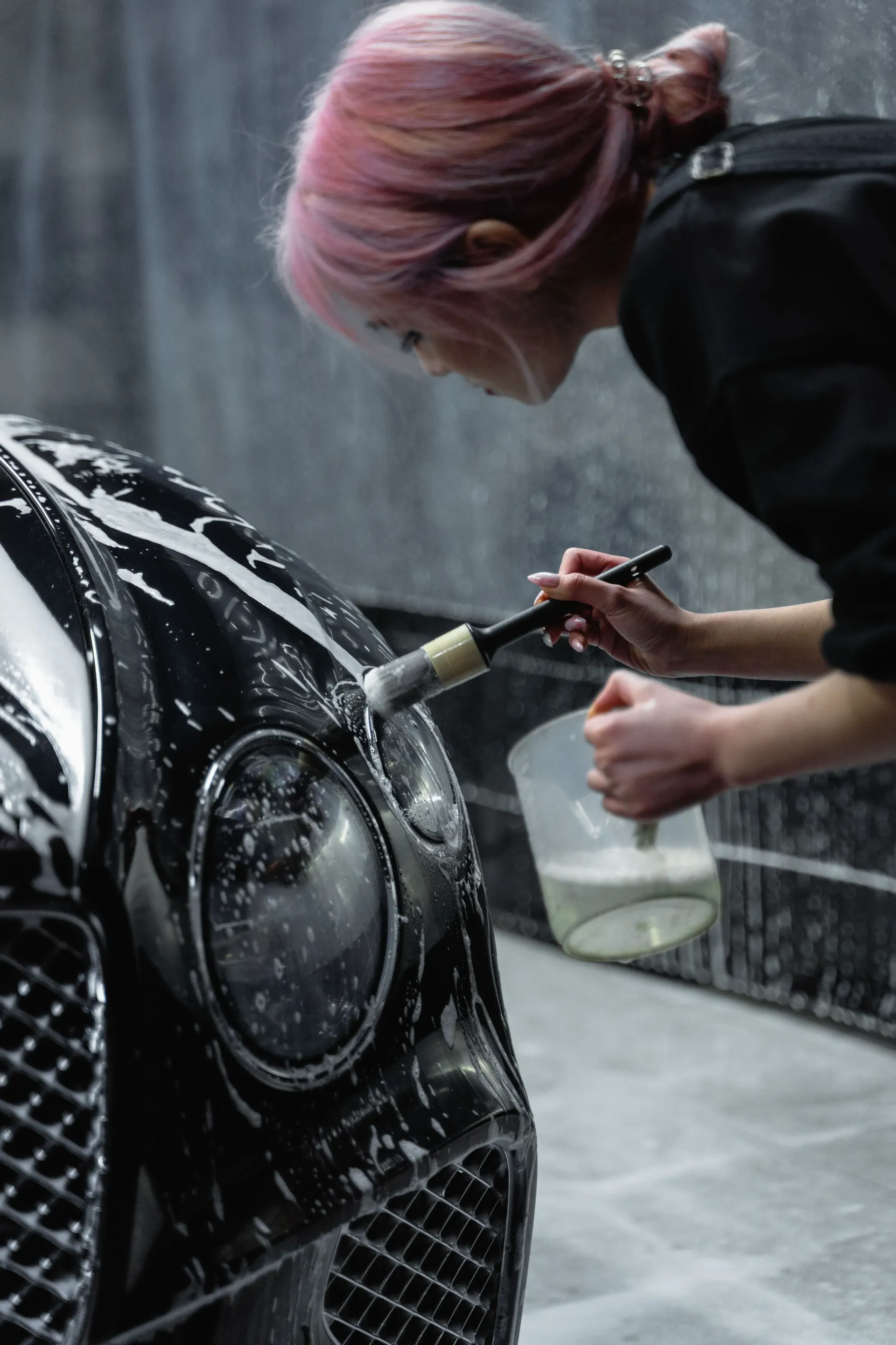 A woman with pink hair uses a soft detailing brush and soapy water to clean the front headlight and grille of a sleek, black luxury sports car.