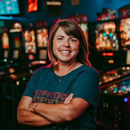 A woman is standing in front of pinball machines with her arms crossed and smiling.