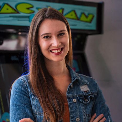 A woman in a denim jacket is smiling in front of a pac man machine