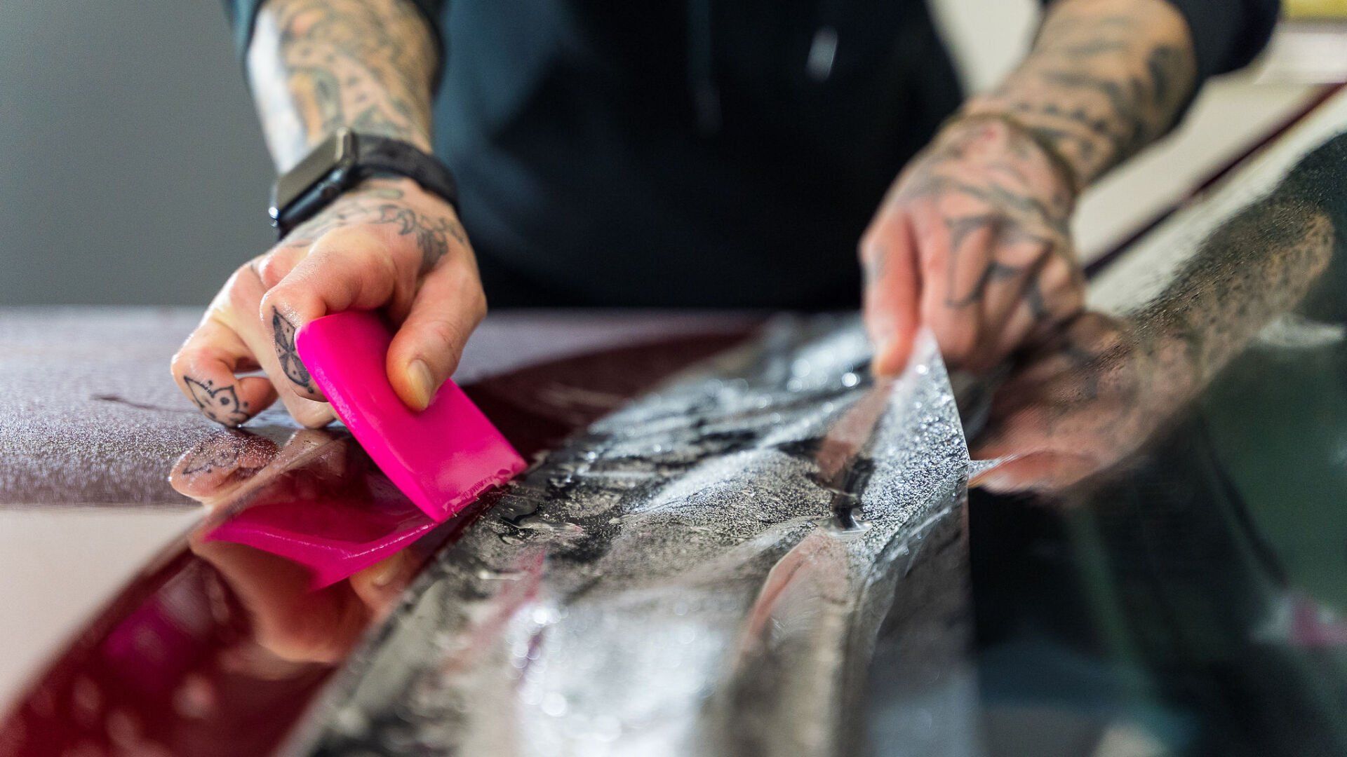 A man with tattoos is wrapping a car with a pink squeegee.