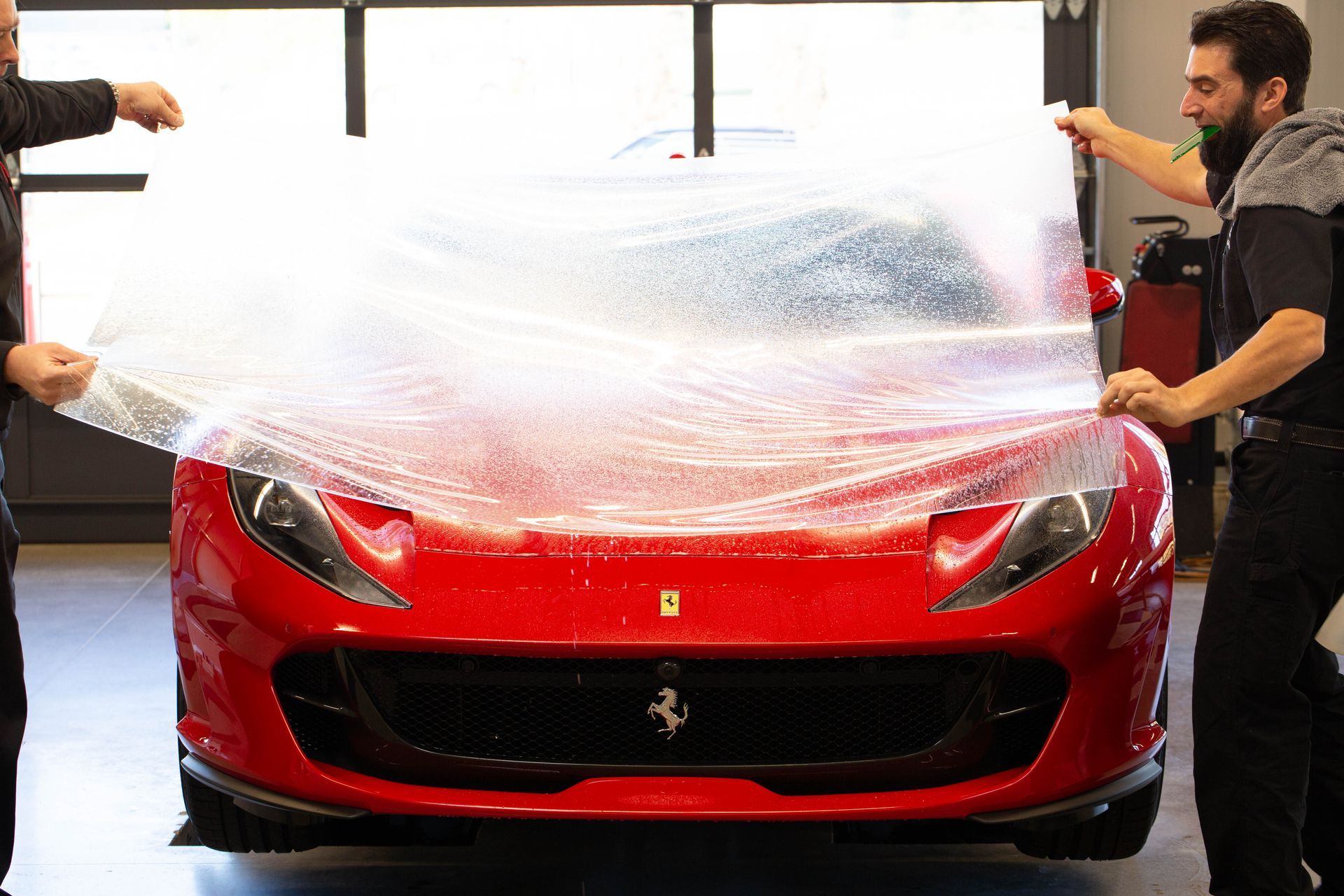 Two people applying clear protective film to a red Ferrari sports car's hood in a shop.