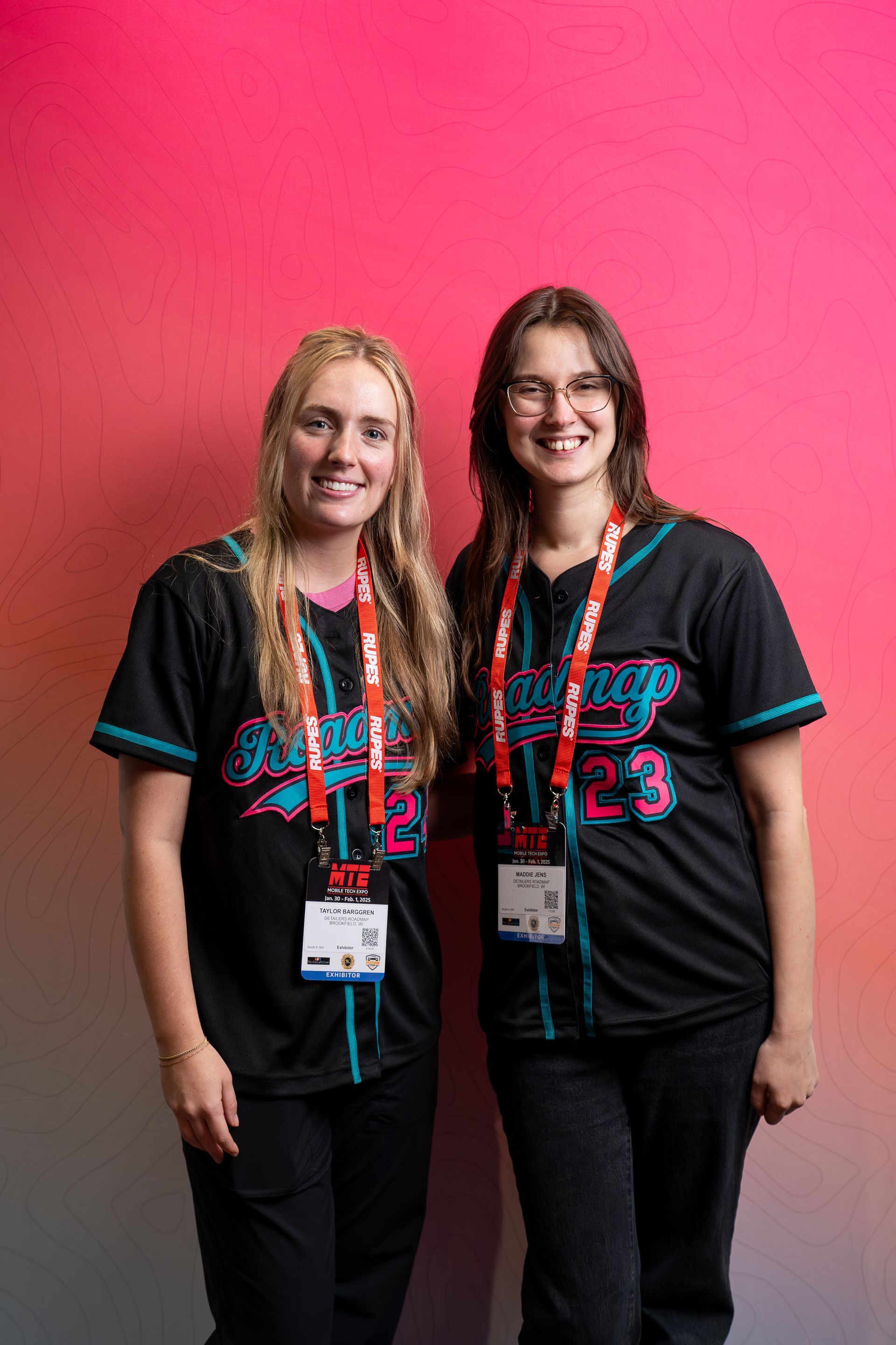 Two women smiling, wearing black jerseys with teal trim, standing in front of a pink and white wall.