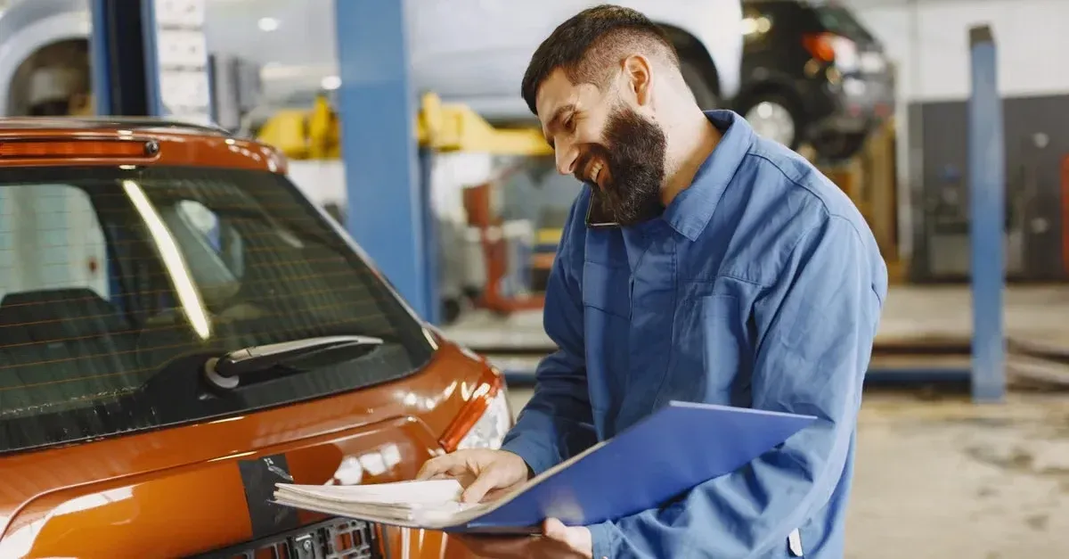Mechanic in blue uniform inspecting orange car engine with clipboard at automotive service facility.