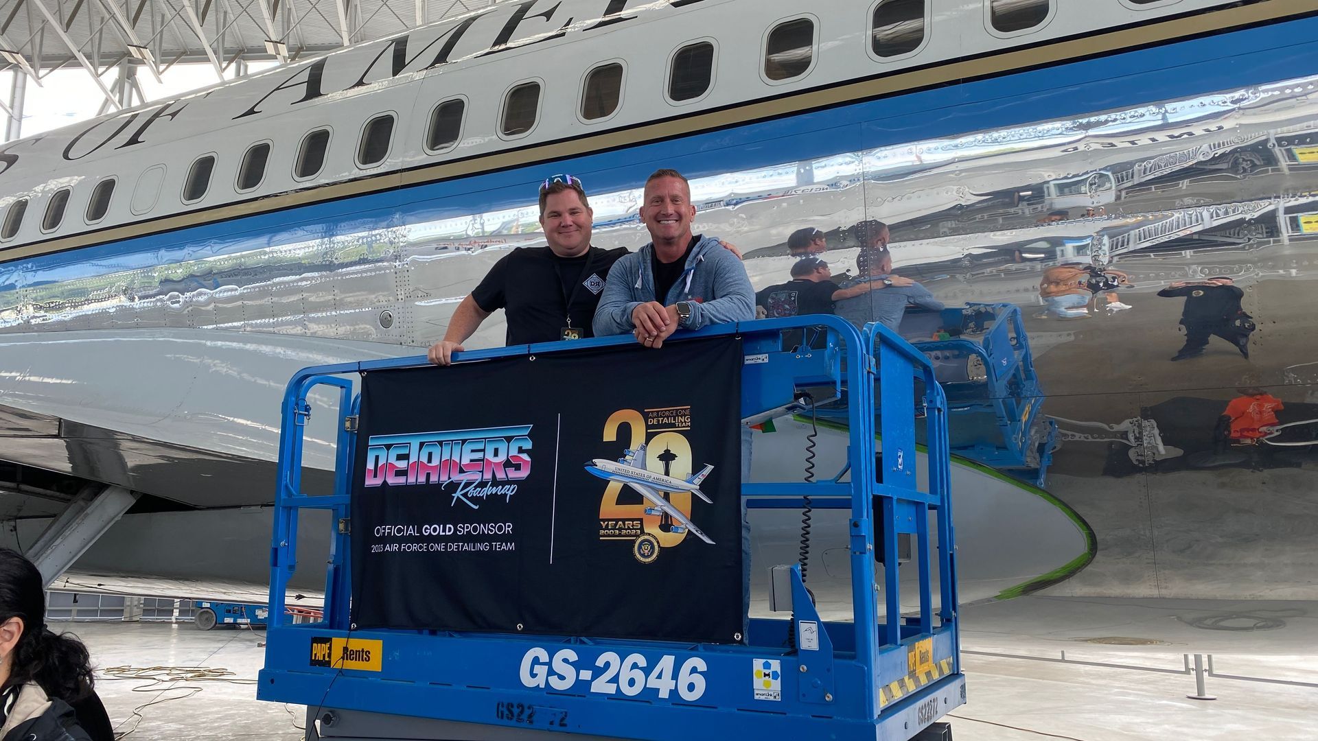 Two men are standing on a lift in front of an airplane.