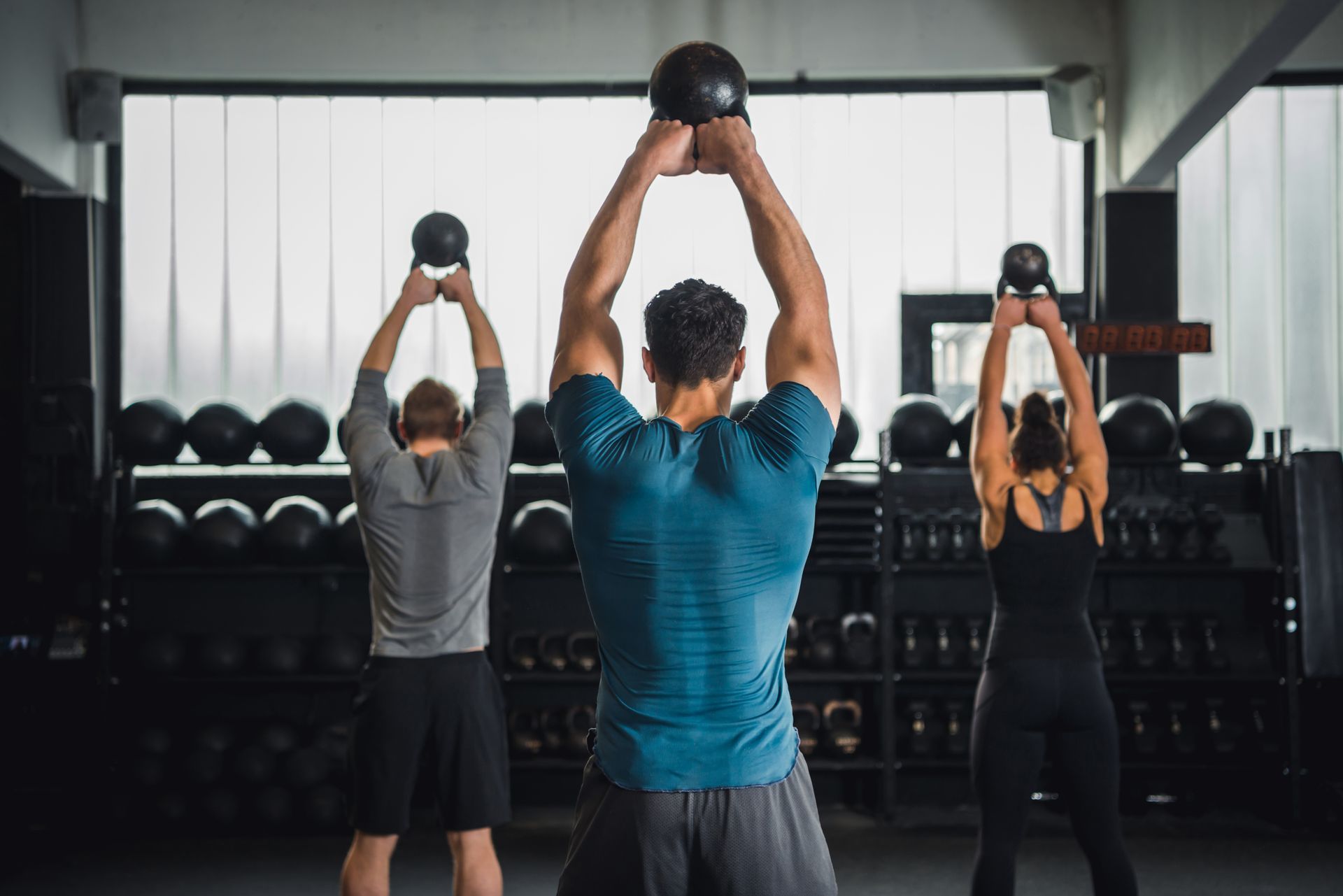 Three people doing overhead kettlebell exercises in a gym.