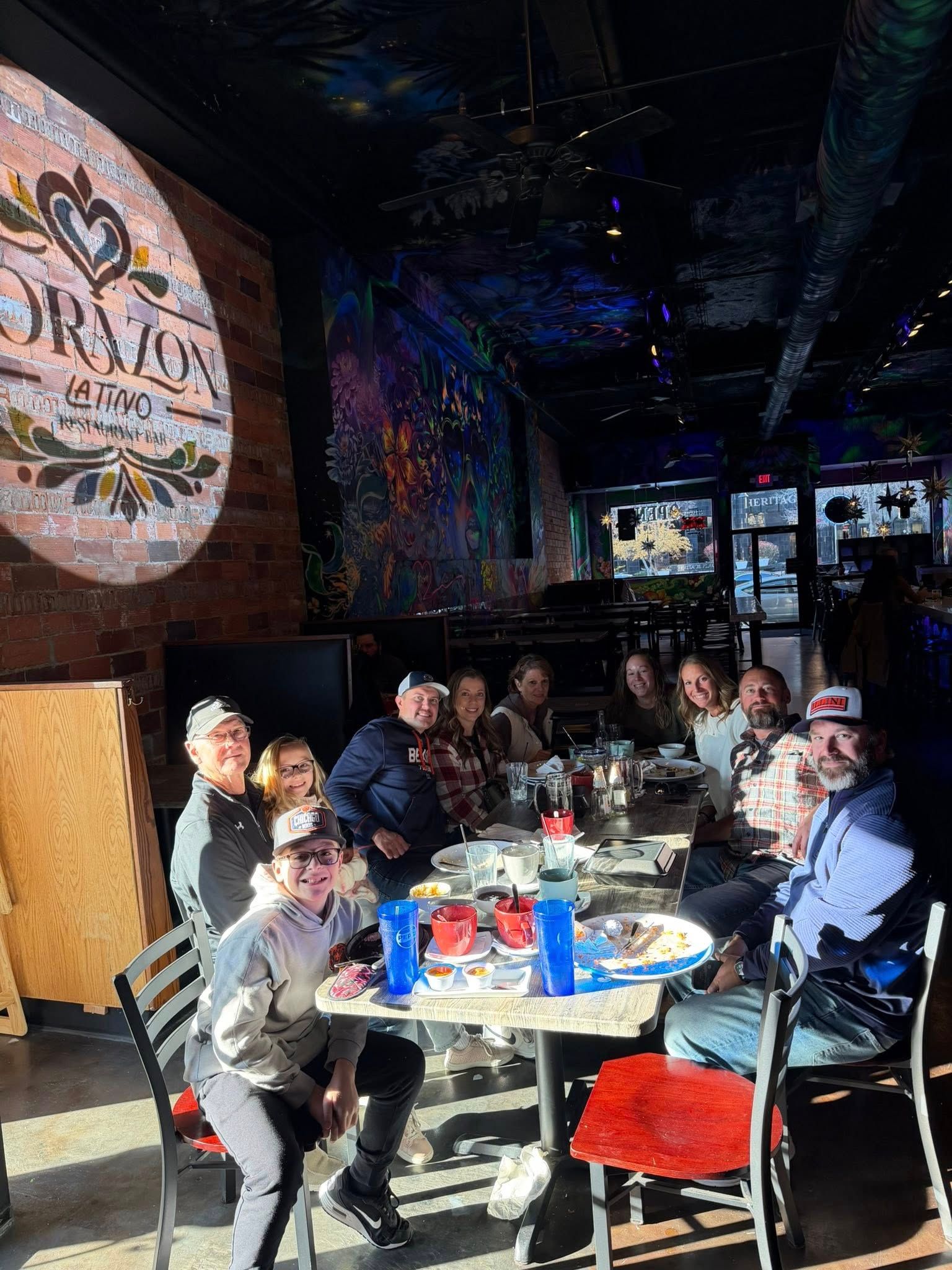 Group of people at a restaurant, sitting at a table with drinks and food. Mural and sunny lighting.