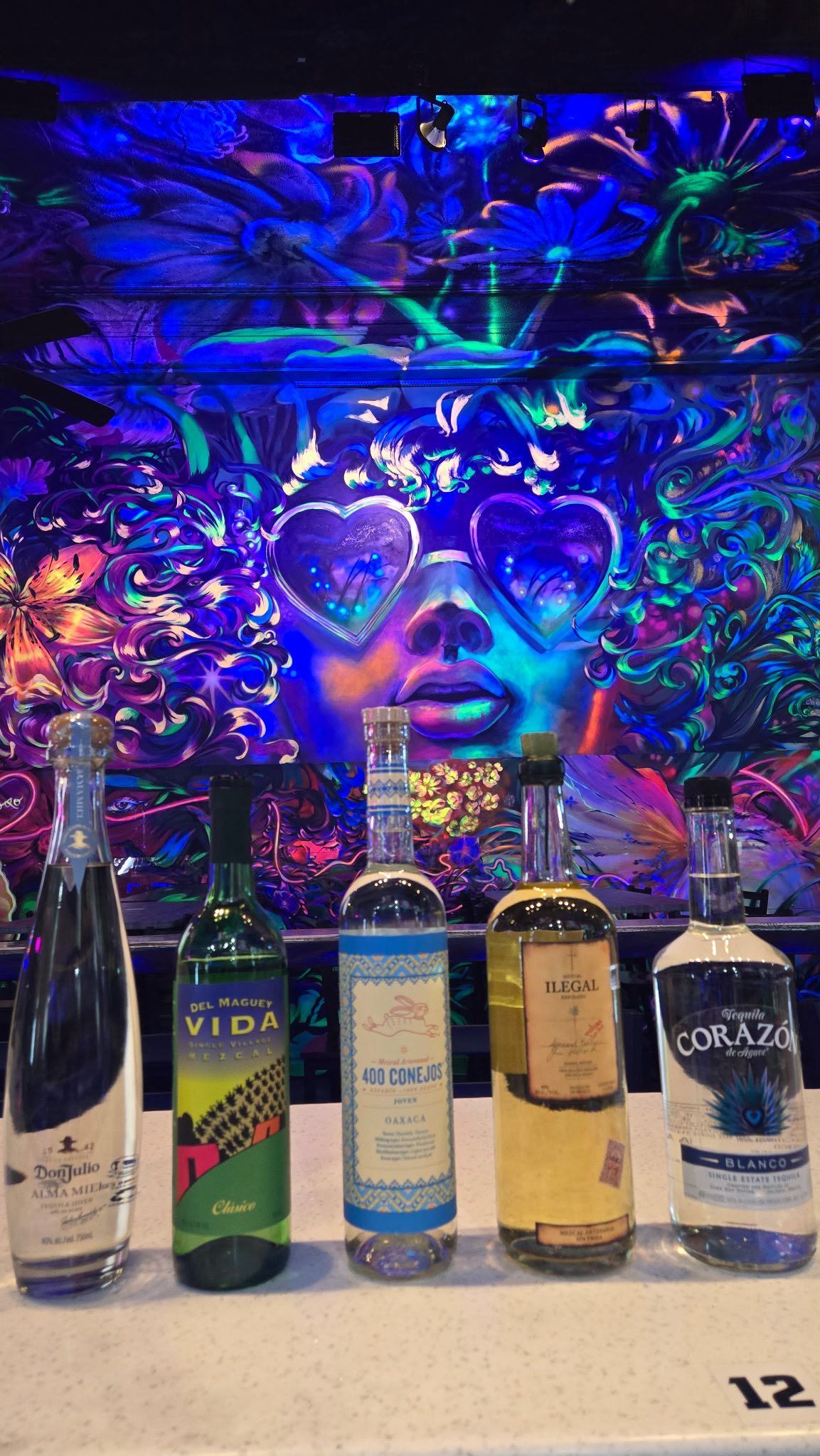 Five liquor bottles in front of a colorful, glowing mural of a face with heart-shaped glasses.