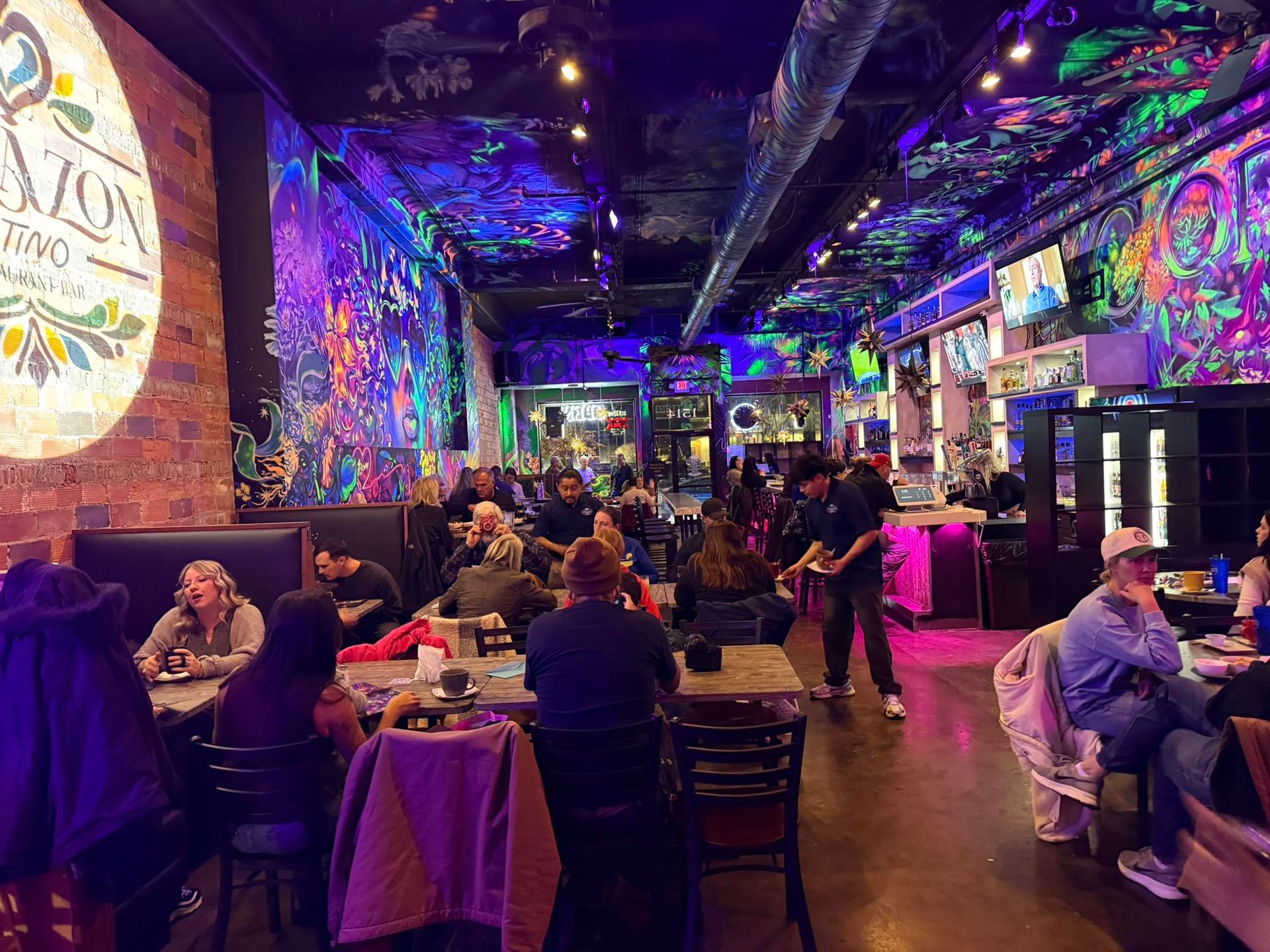 Interior of a busy restaurant with colorful murals. People seated at tables, a server walks by.